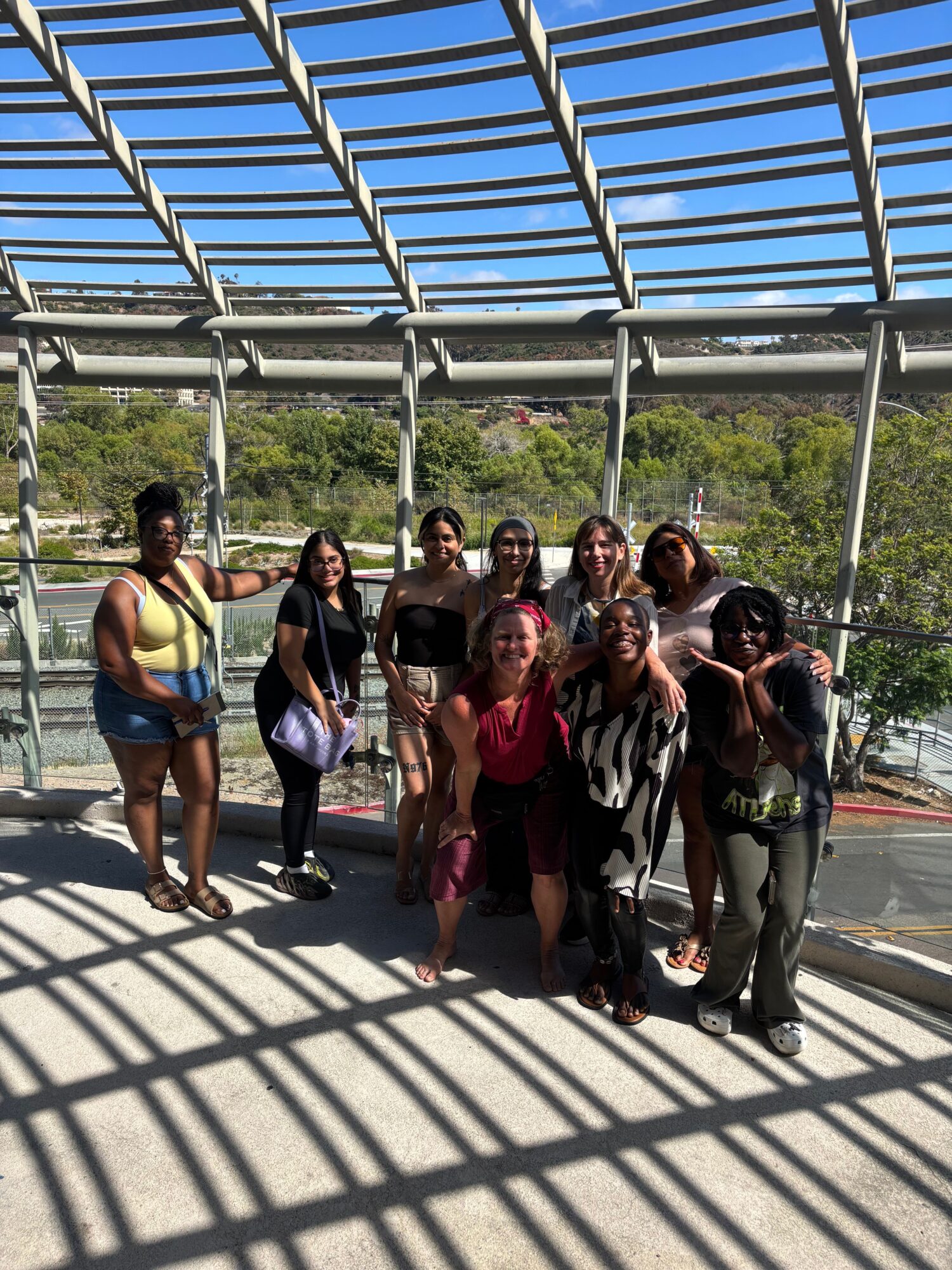 Group of people standing on a bridge with metal railings, shadows cast on the ground, trees and sky in background.