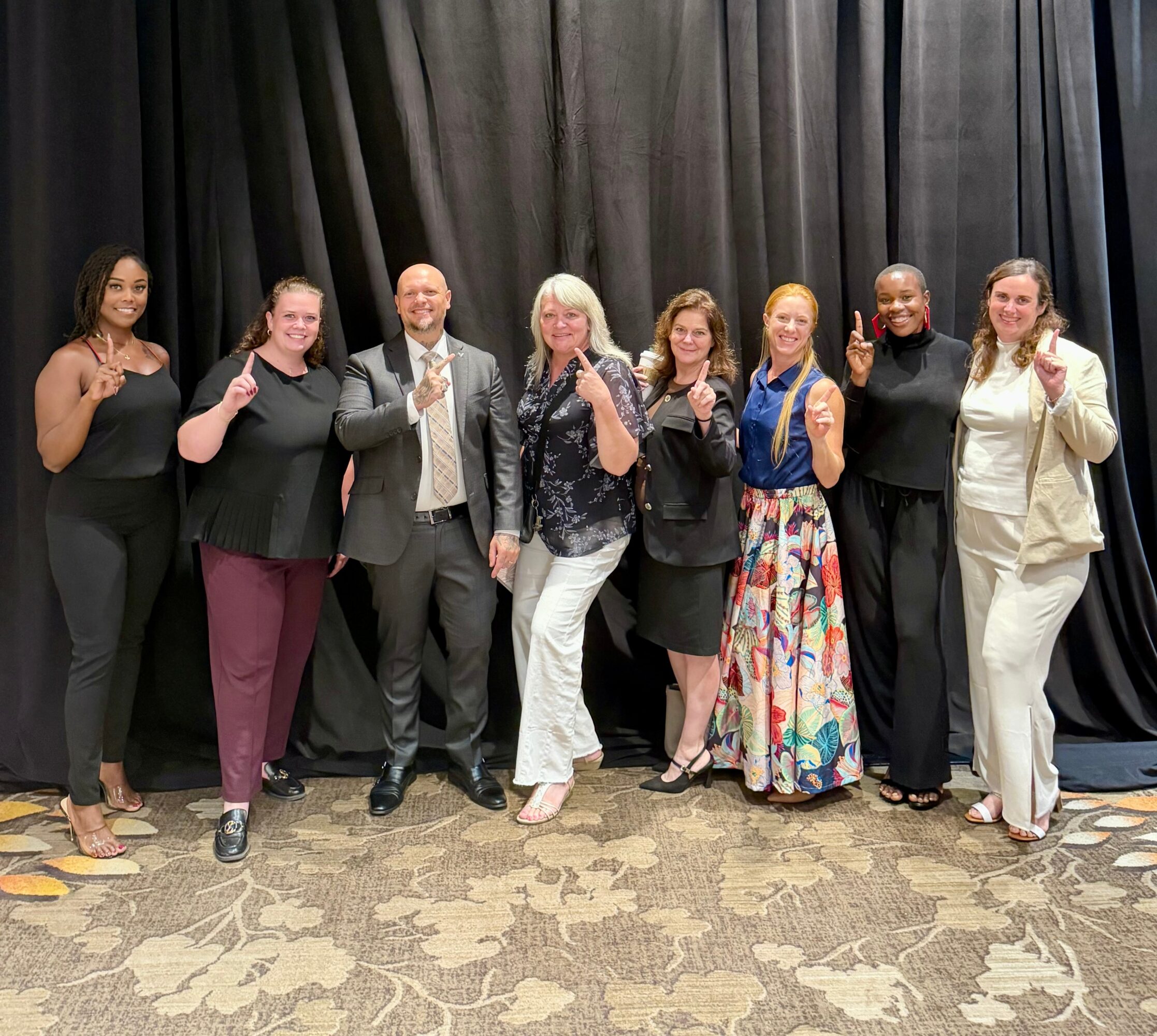 Group of nine diverse women standing on stage in front of black curtains, smiling and making peace signs.