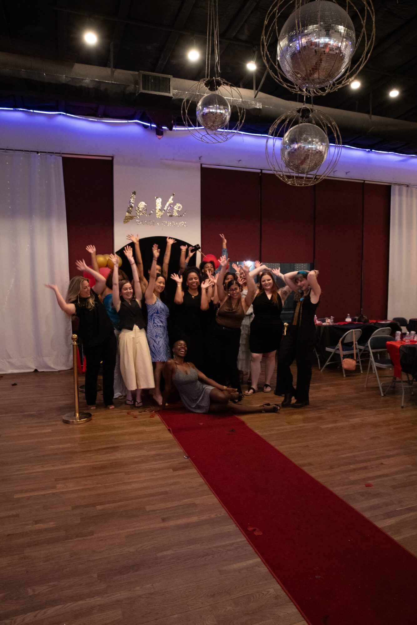 Group of people posing on stage with a decorated backdrop, some raising hands, in a decorated event space with hanging disco balls.