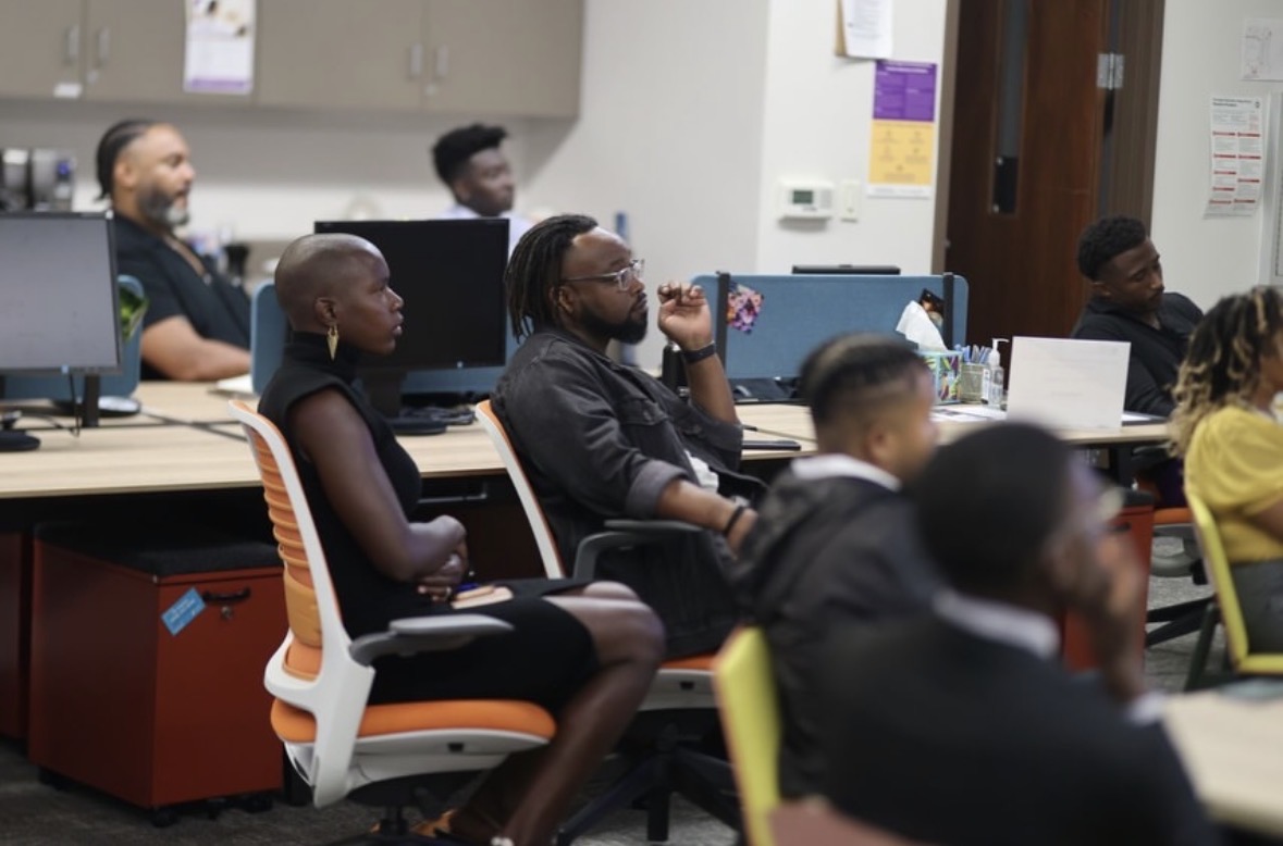 Group of people seated in a classroom or training room, some using computers, listening attentively.