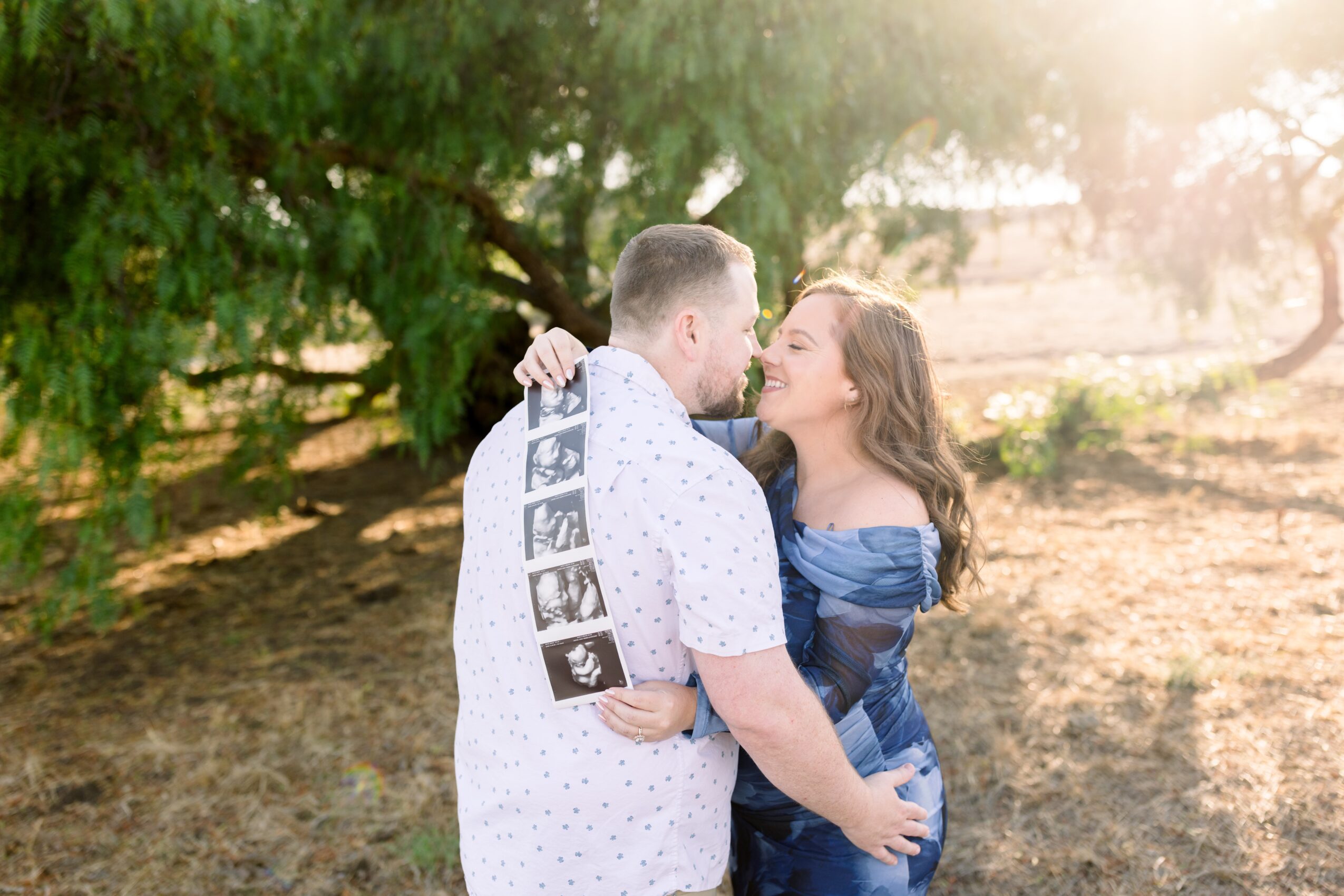 A couple stands close together outdoors, about to kiss, with trees and sunlight in the background.