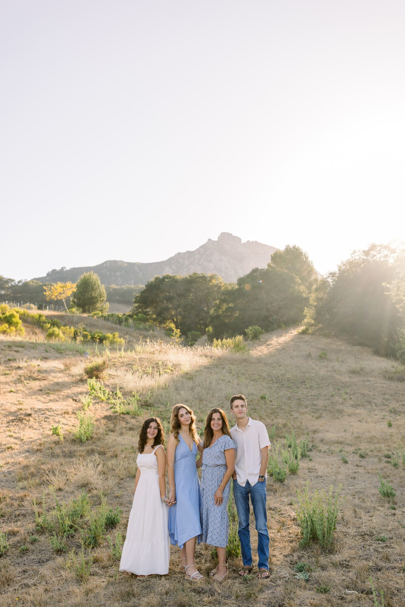 Four people standing outdoors in a grassy field with hills and trees in the background, bright sunlight overhead.