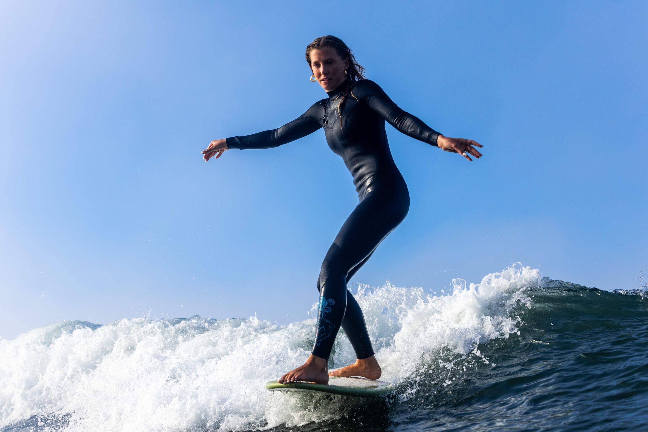 Woman in wetsuit surfing on a wave against a blue sky, arms outstretched for balance.