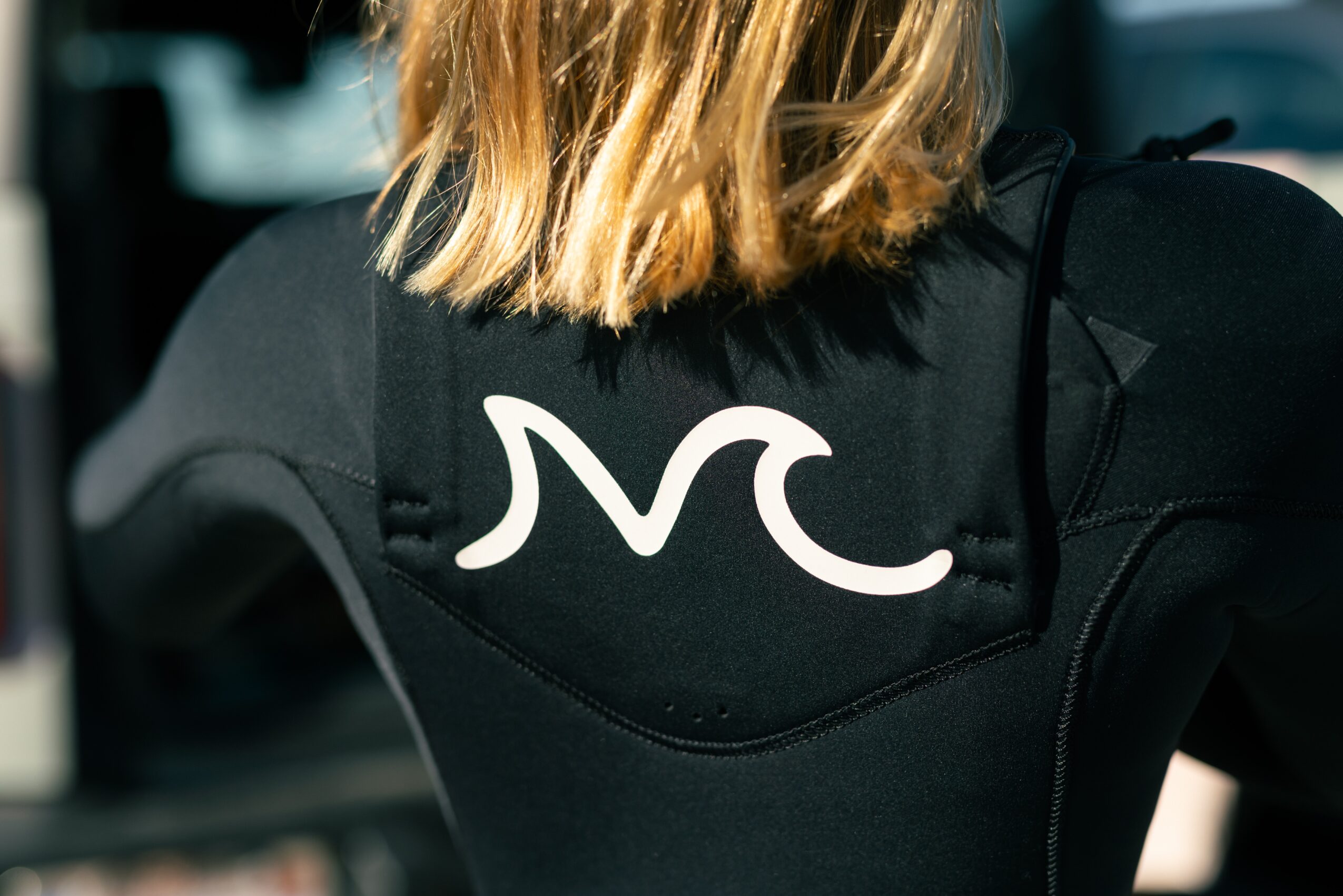 Back of a person wearing a black jacket with a white logo, shoulder-length blonde hair, outdoors.