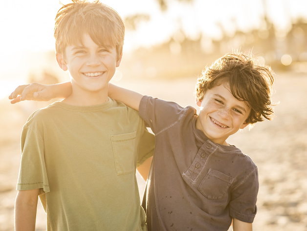 Two boys smiling and standing outdoors with sunlight behind them, one with arm around the other's shoulder.