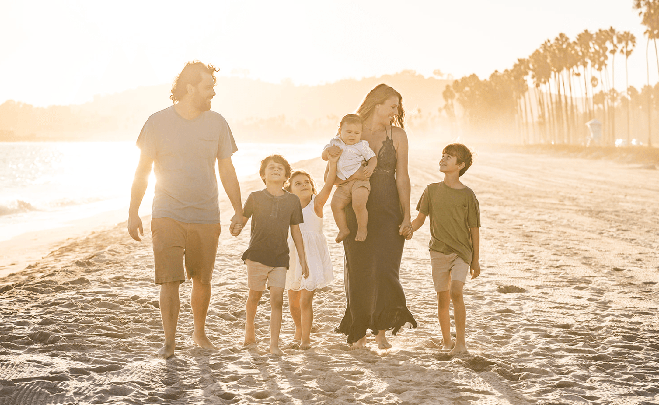 Group of seven people walking on a beach at sunset, including adults and children, with trees in the background.