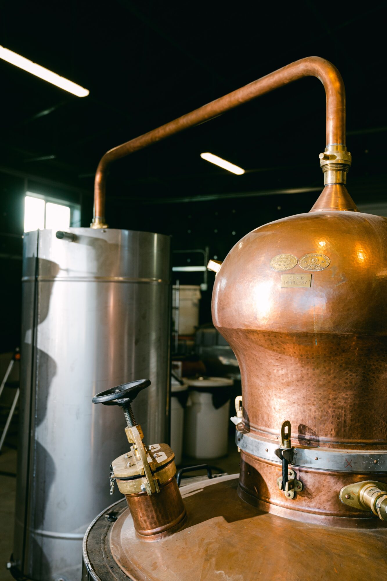 Copper distillation apparatus with a large round vessel connected to a vertical metal tank, in a distillery setting.