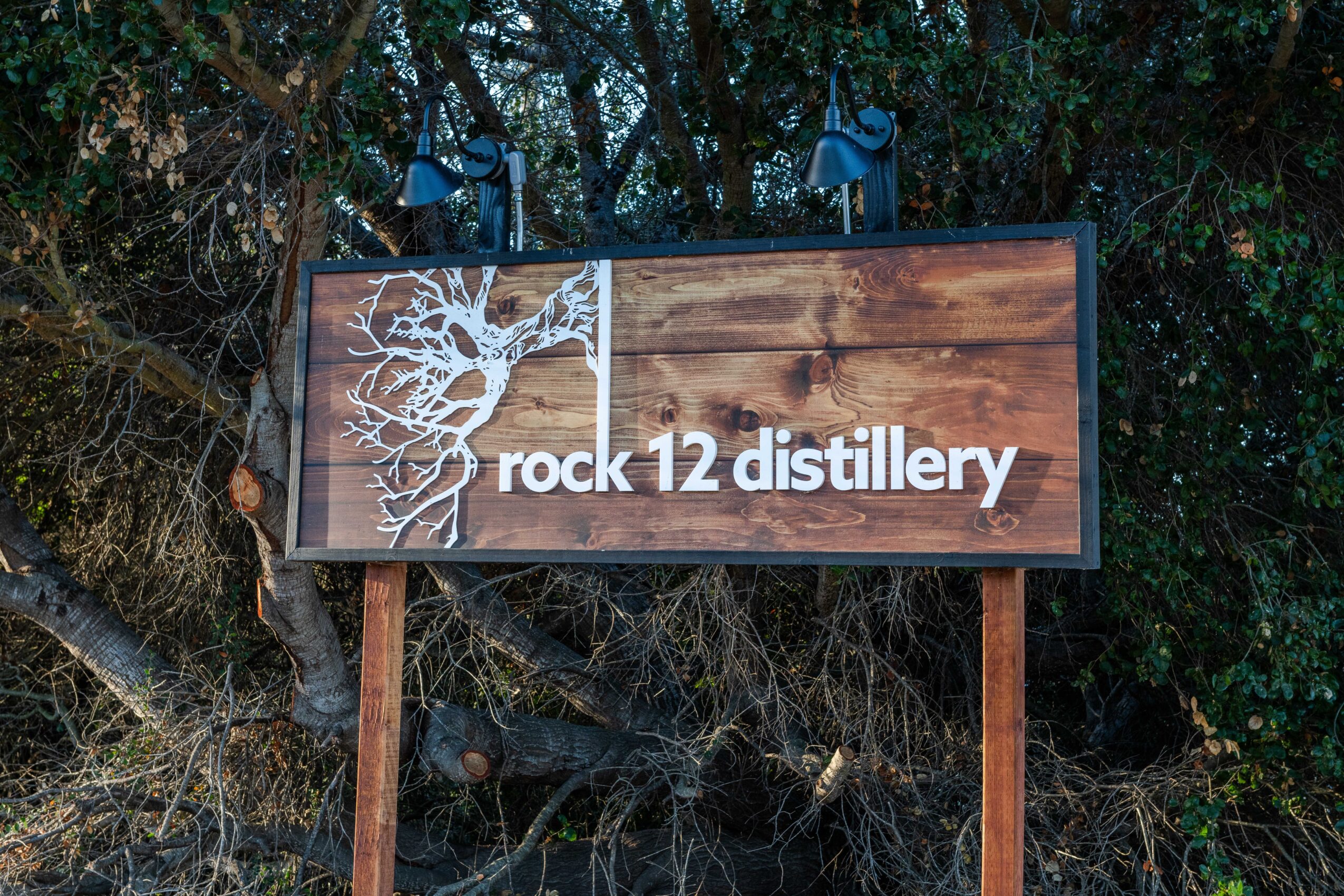 Wooden sign with tree graphic and text reading 'rock 12 distillery' against a background of trees and bushes.