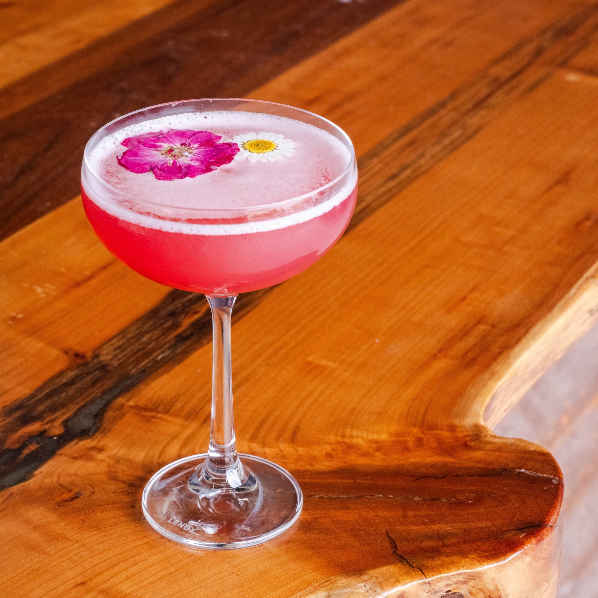 Pink cocktail with flower garnish in a glass on a wooden surface.