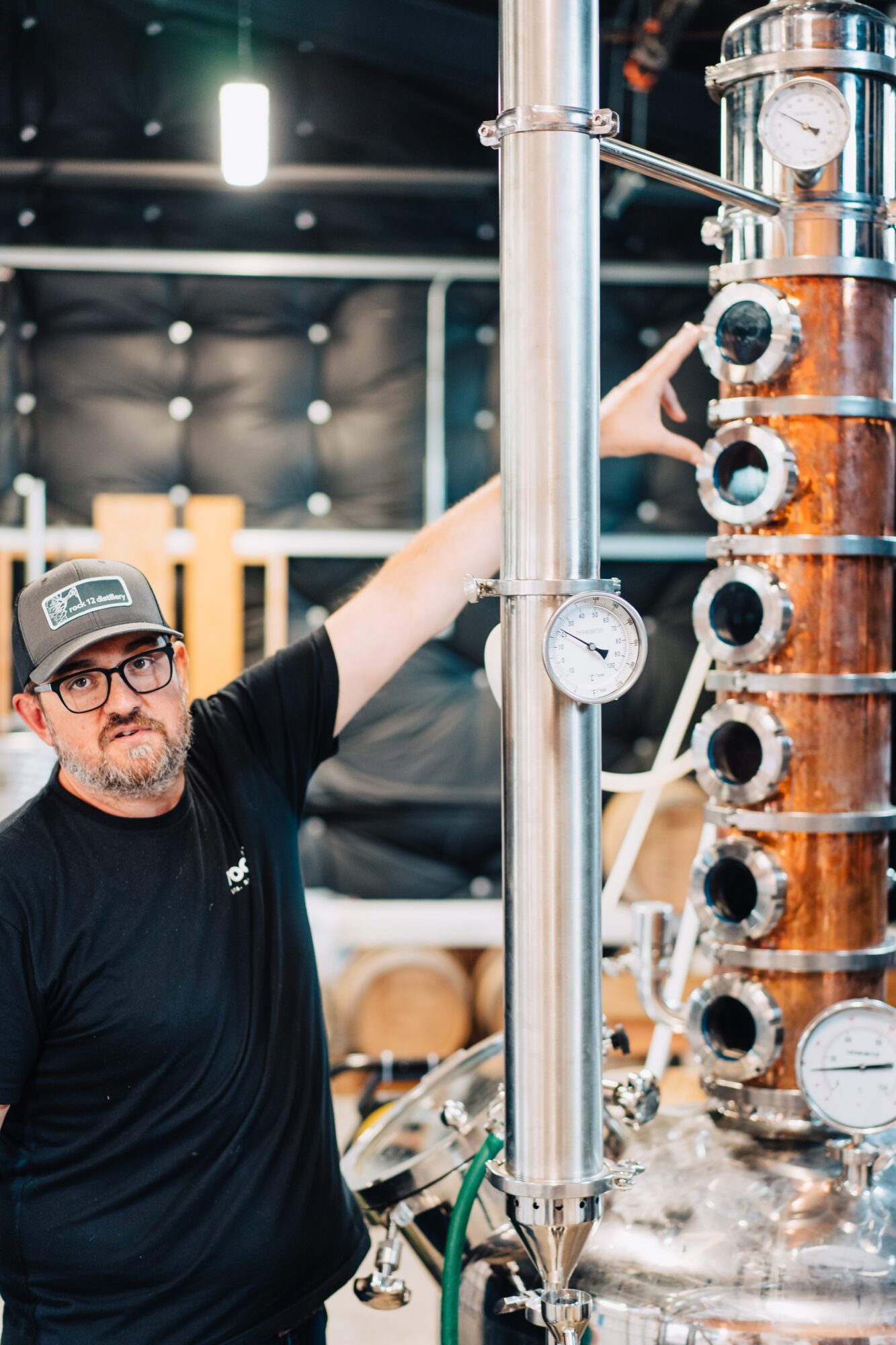 Man with glasses and a cap operating a distillation apparatus with gauges and pipes in a brewery or distillery setting.