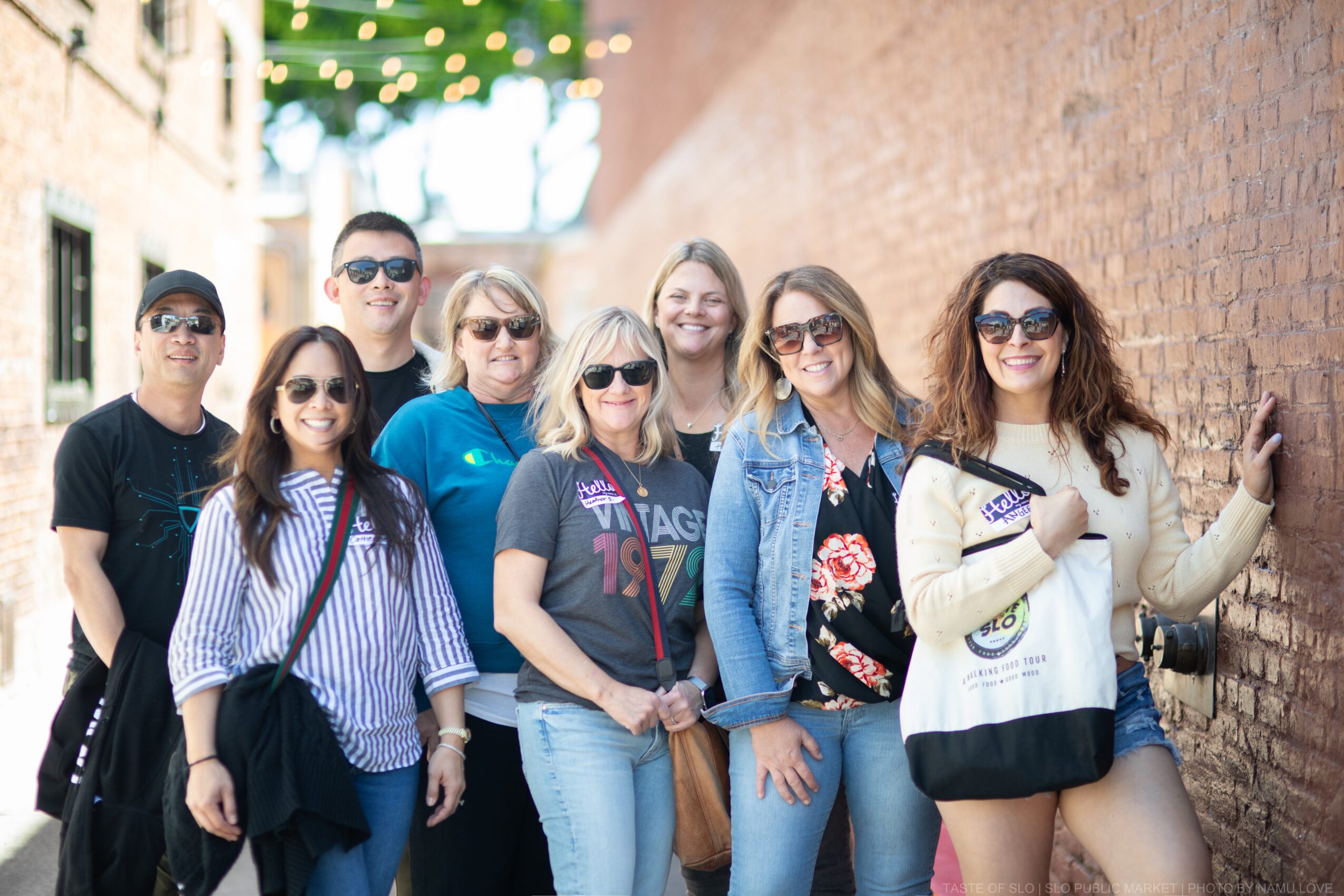 Group of eight people standing outdoors against a brick wall, smiling, wearing casual clothing and sunglasses.