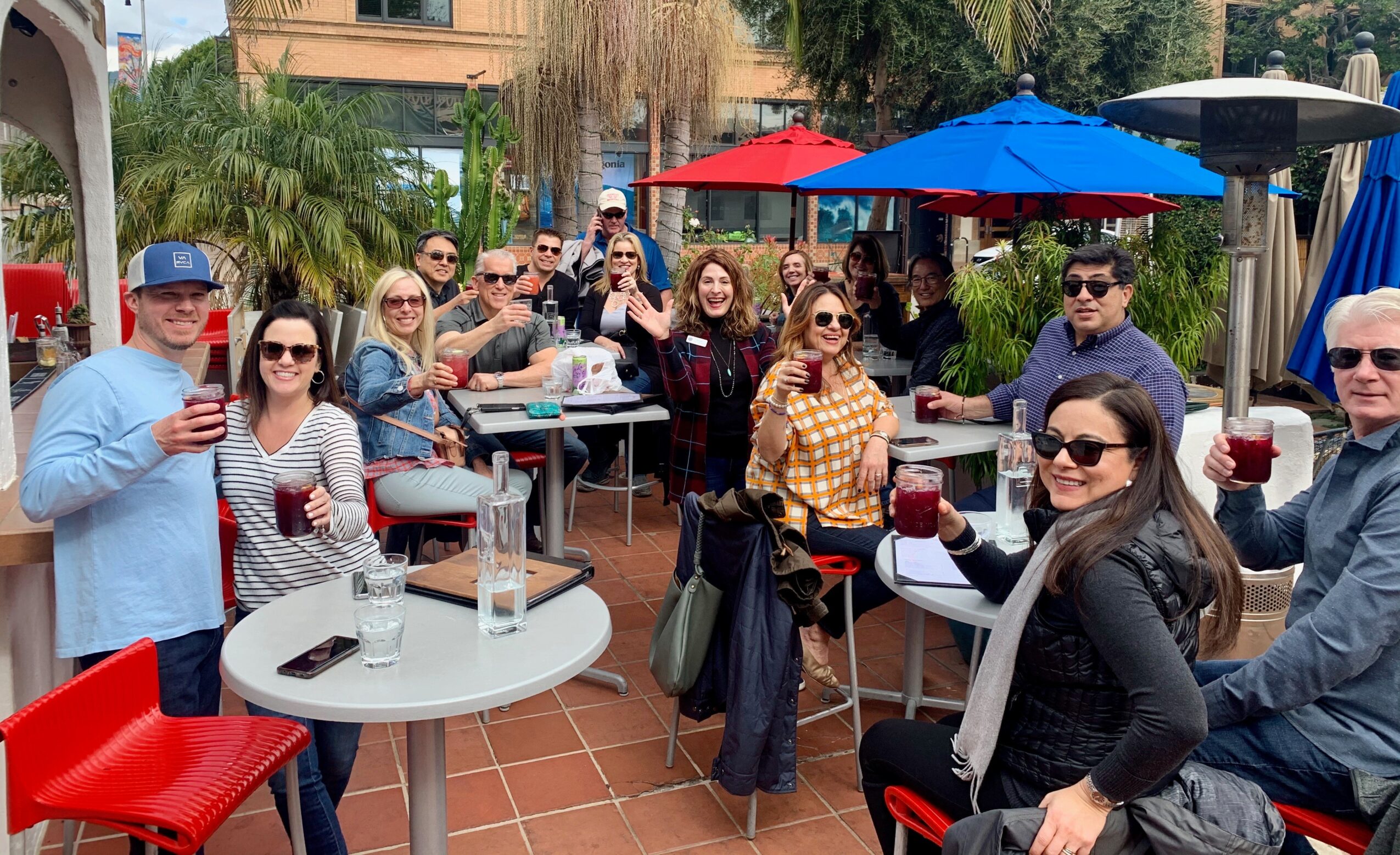 Group of people enjoying drinks at an outdoor patio with umbrellas and tables, smiling and raising glasses.