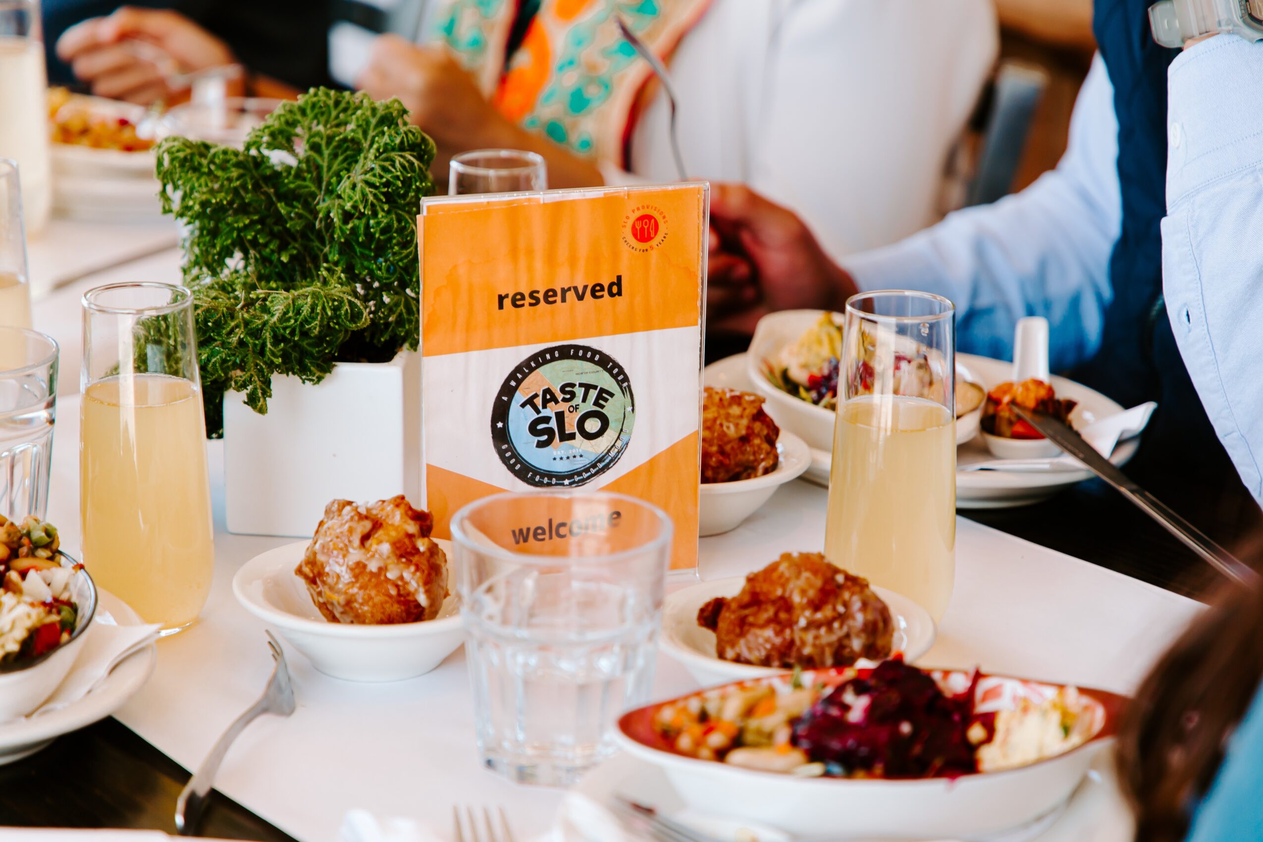 Table with food, drinks, and a sign reading 'Taste SLO' surrounded by people dining.