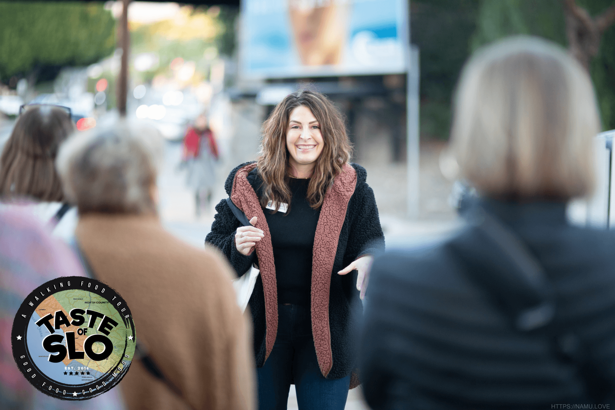 Smiling woman with long hair standing outdoors, surrounded by people, with a blurred background of trees and a billboard.