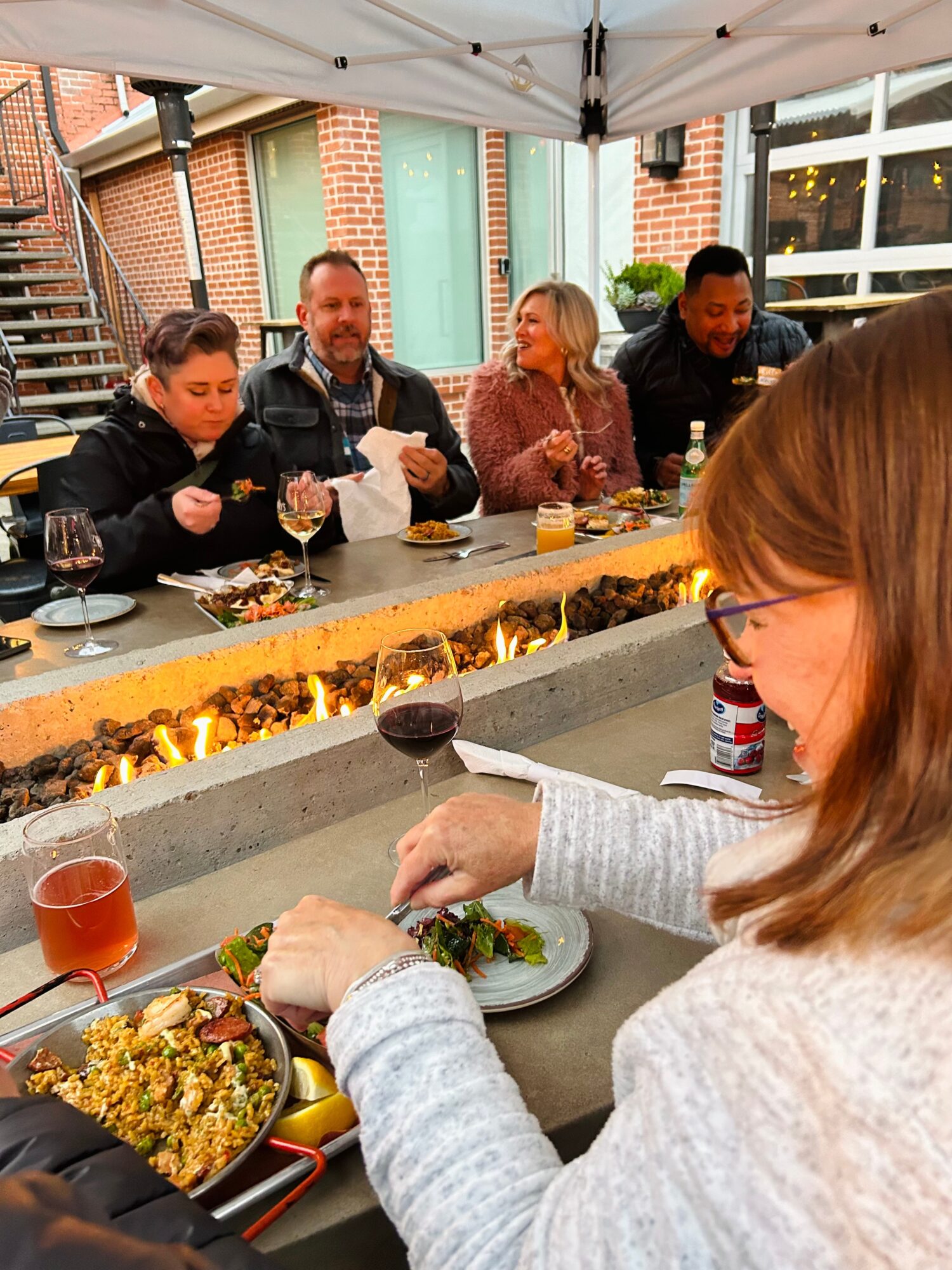 Group of five people sitting at an outdoor bar, enjoying food and drinks under a canopy, with a brick building in the background.