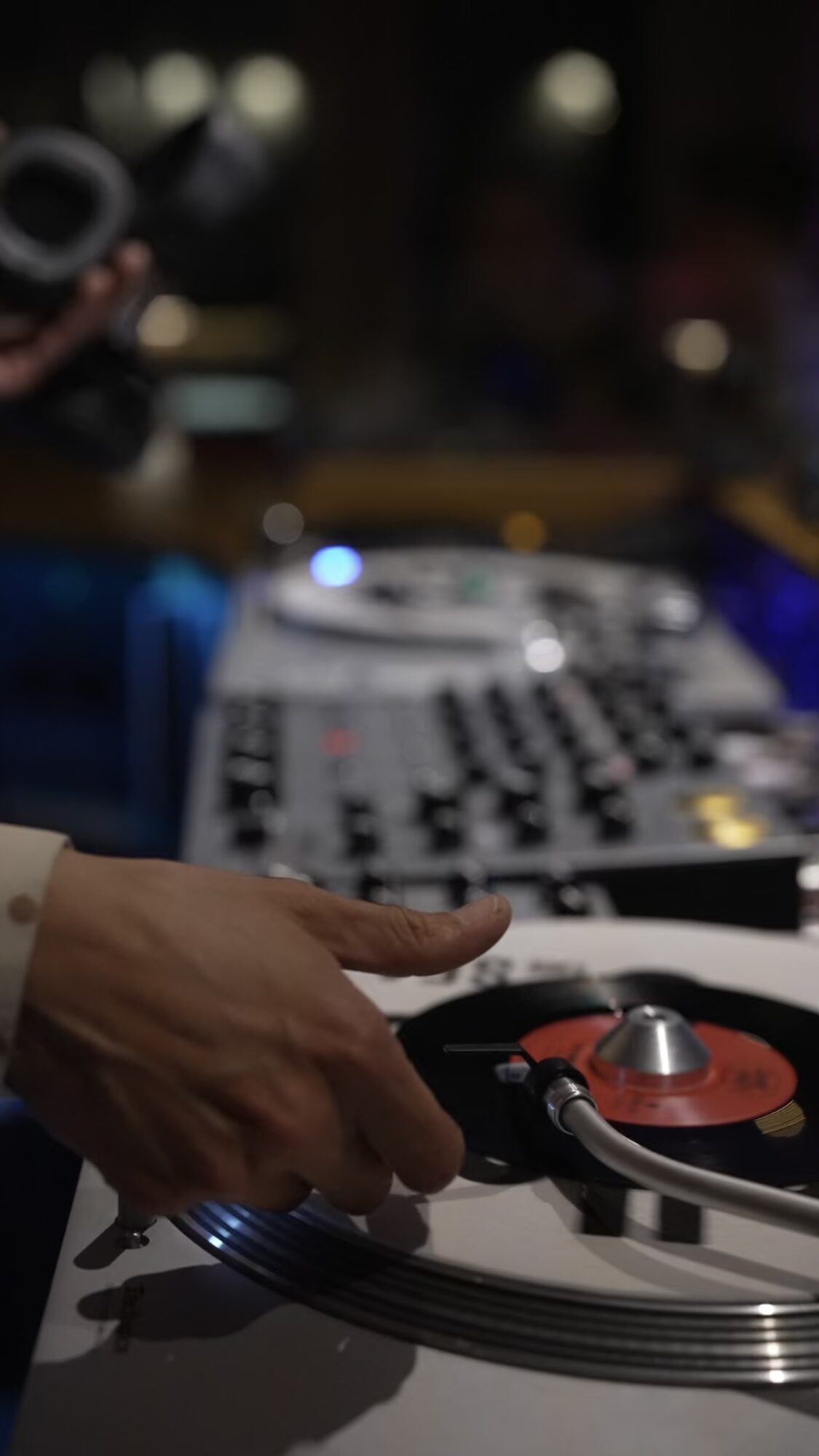 Person's hand adjusting a turntable with a vinyl record, blurred DJ equipment in background, dim lighting, indoor setting.