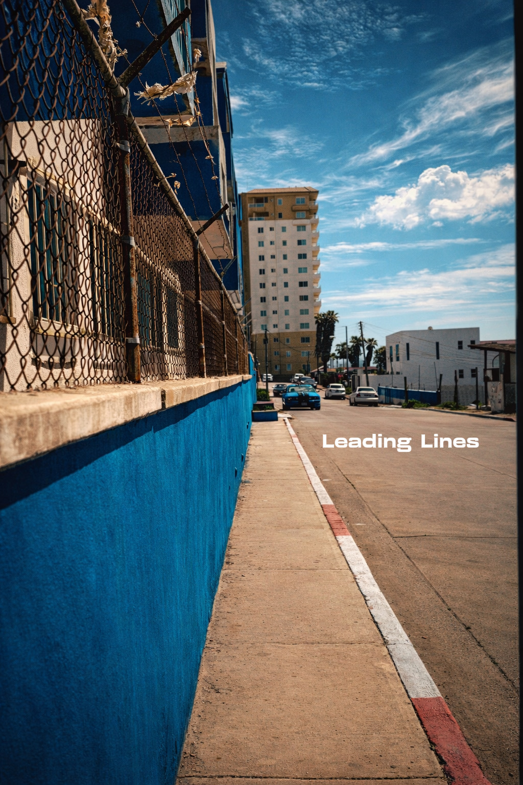 Sidewalk with a blue wall, leading to a street with buildings and parked cars, under a partly cloudy sky.