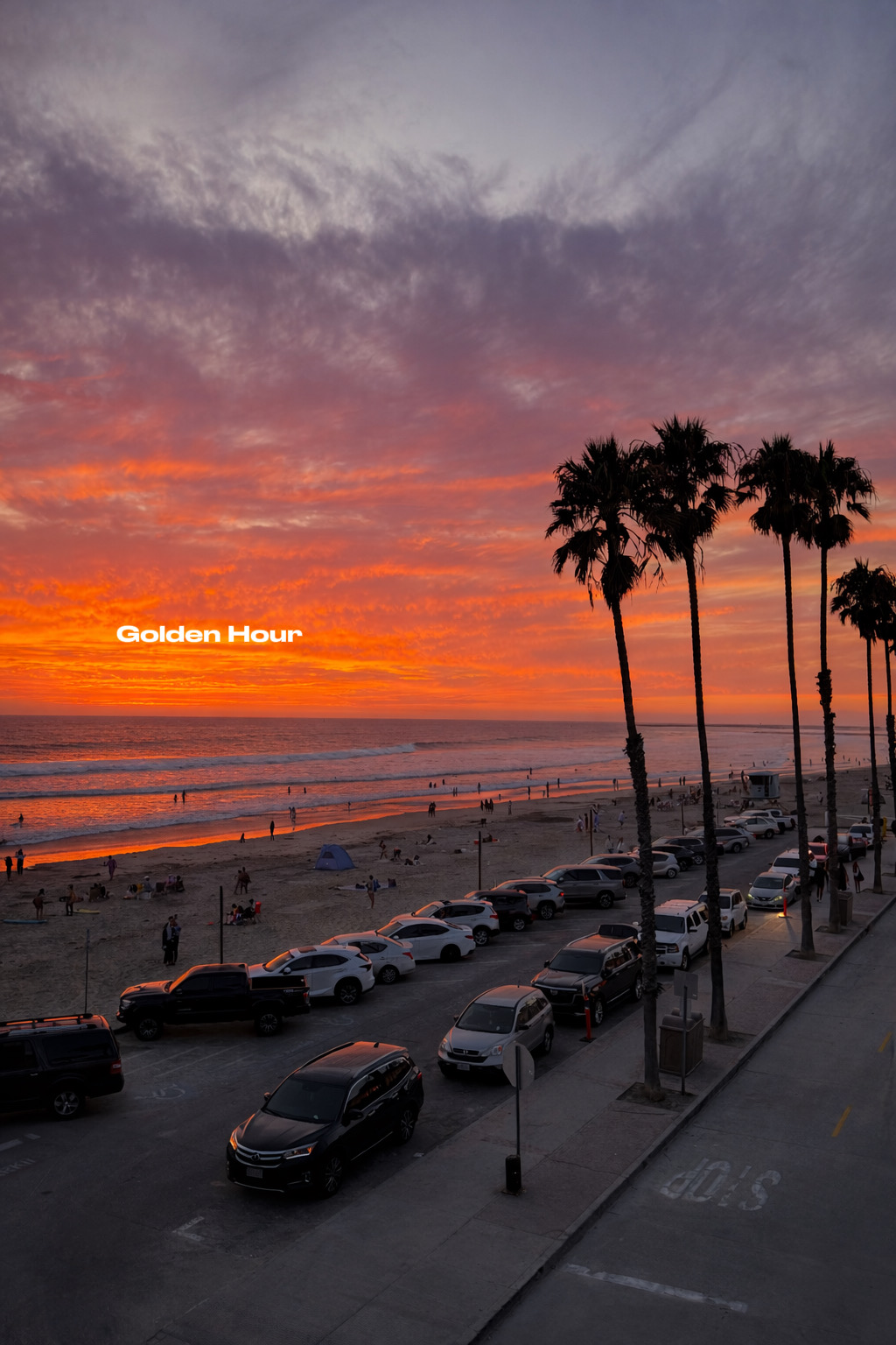 Sunset over a beach with palm trees and parked cars along the street, colorful sky with clouds, and people on the beach.