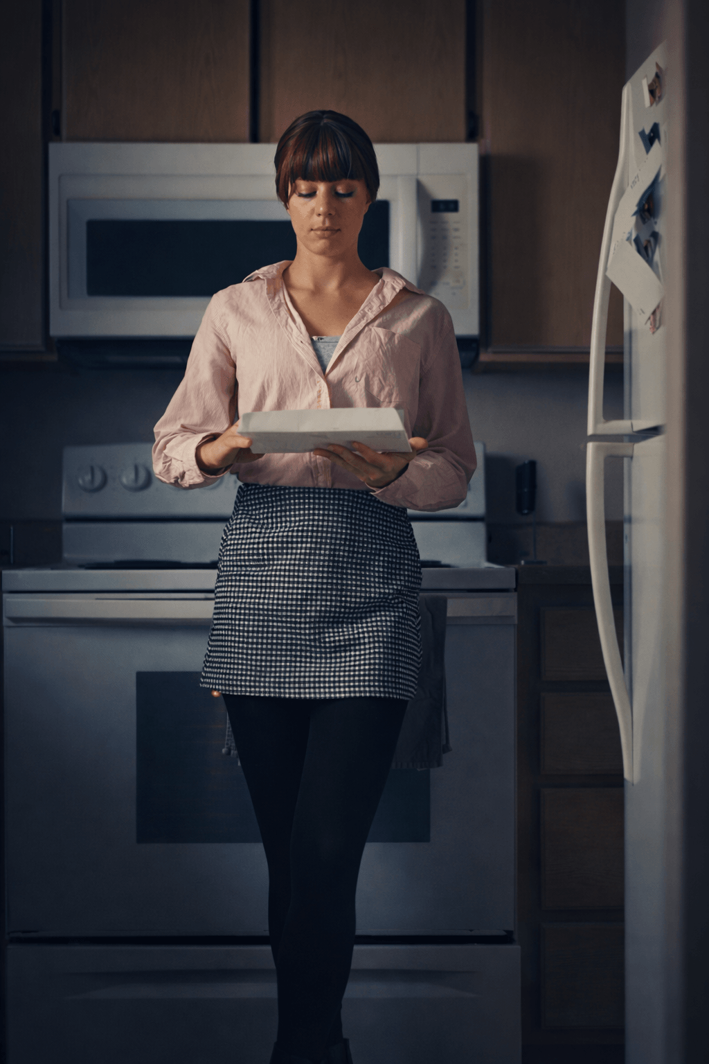 Woman standing in kitchen holding a tray, wearing a pink blouse and checkered skirt, with a microwave and refrigerator behind her.