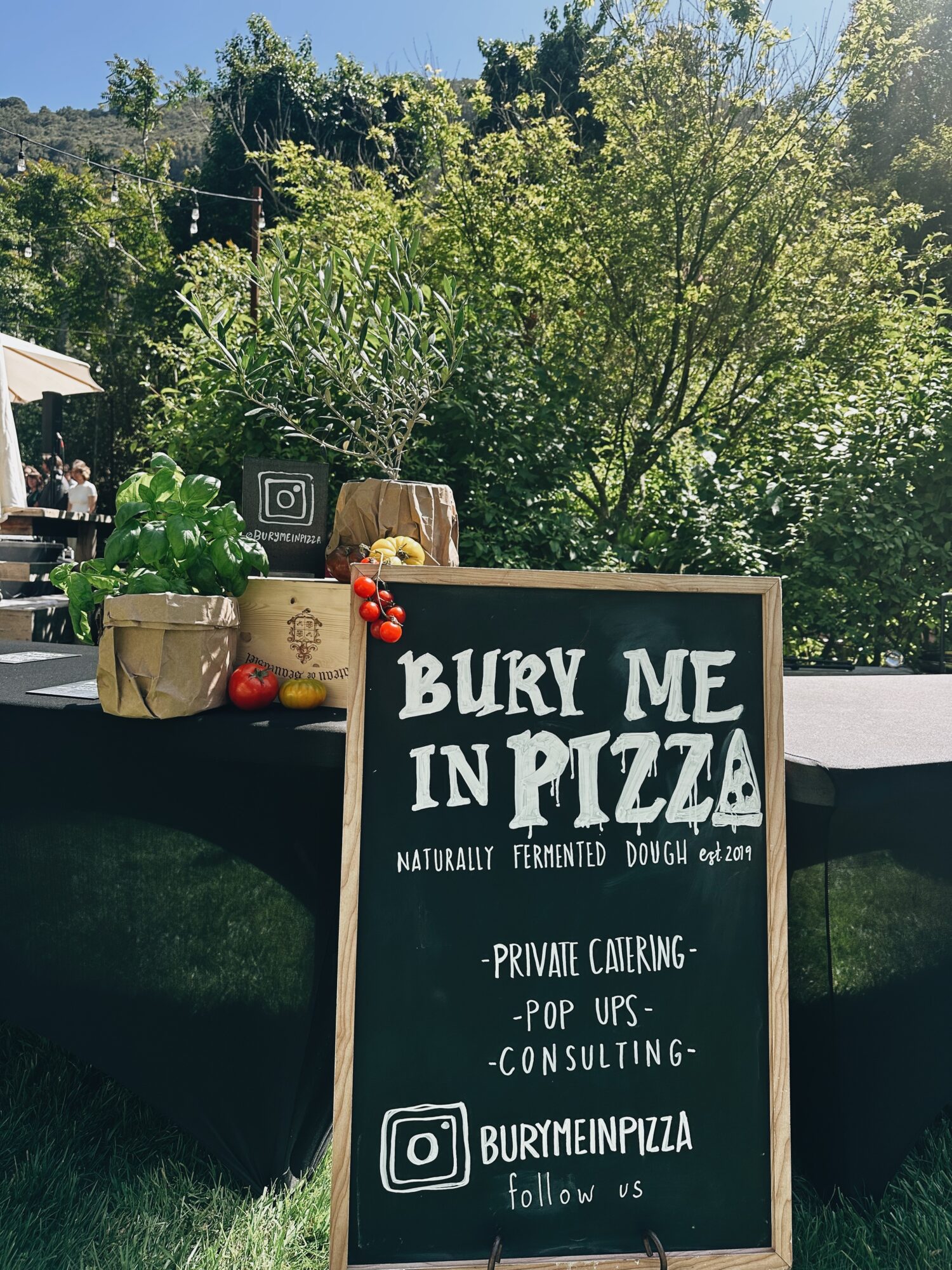 Chalkboard sign advertising pizza with tomatoes and herbs on a table outdoors, surrounded by trees and greenery.
