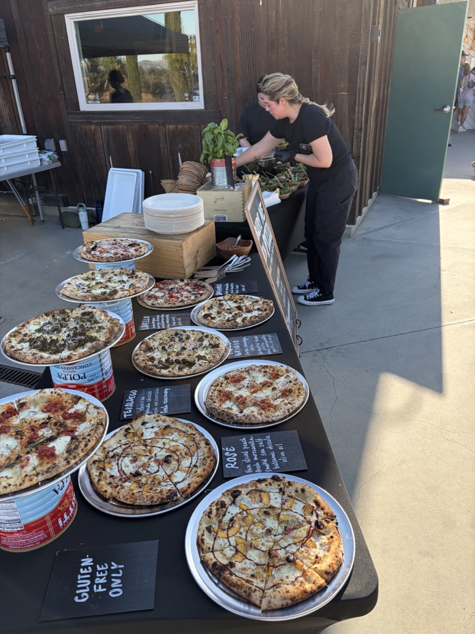 Person serving pizza at outdoor table with various pizzas displayed, wooden wall background, sunny day.