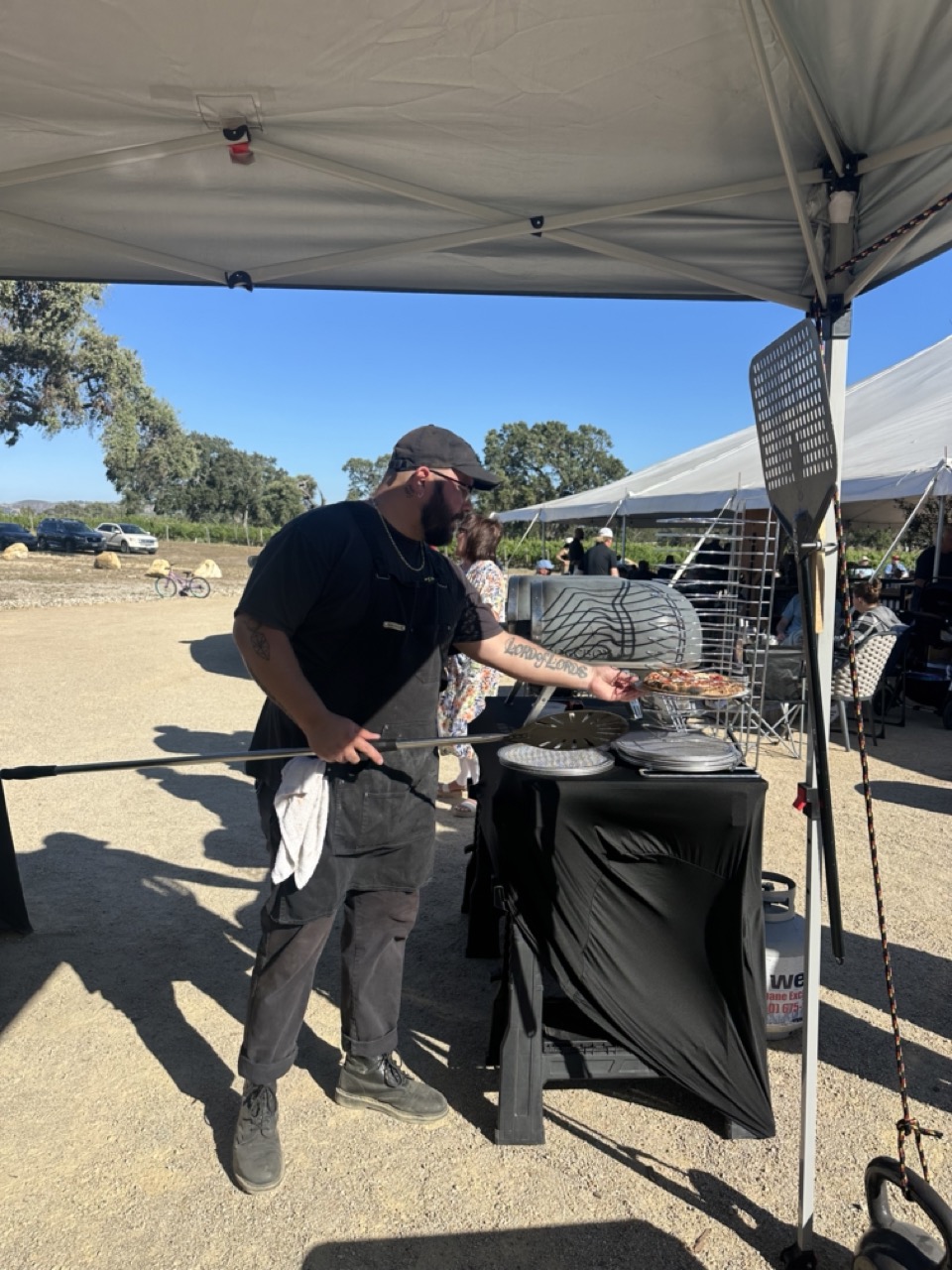 Person cooking on outdoor grill under a canopy, with tents and trees in the background.