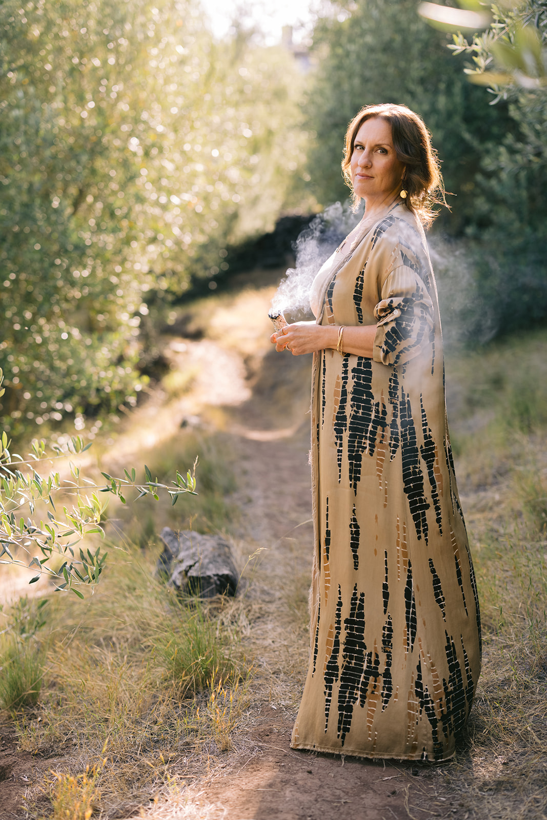 Woman standing outdoors on a grassy path surrounded by trees, wearing a long patterned dress and looking to the side.