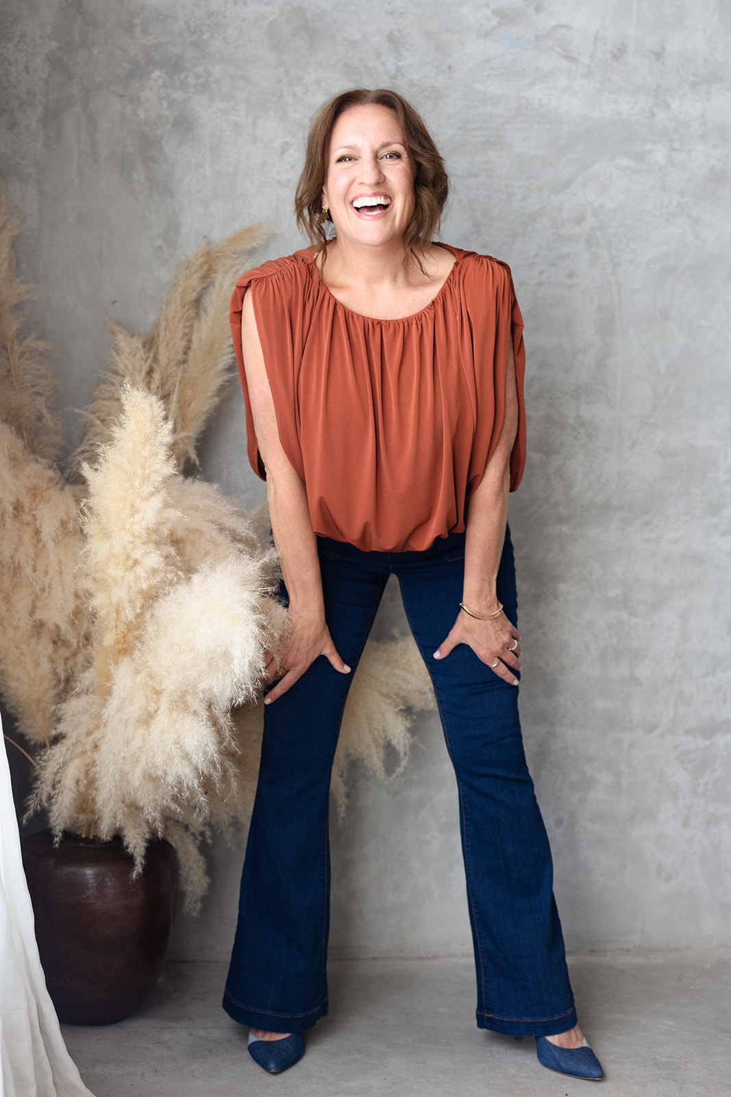 Woman laughing, standing against a textured gray wall with pampas grass in a large pot, wearing a rust-colored top and dark jeans.
