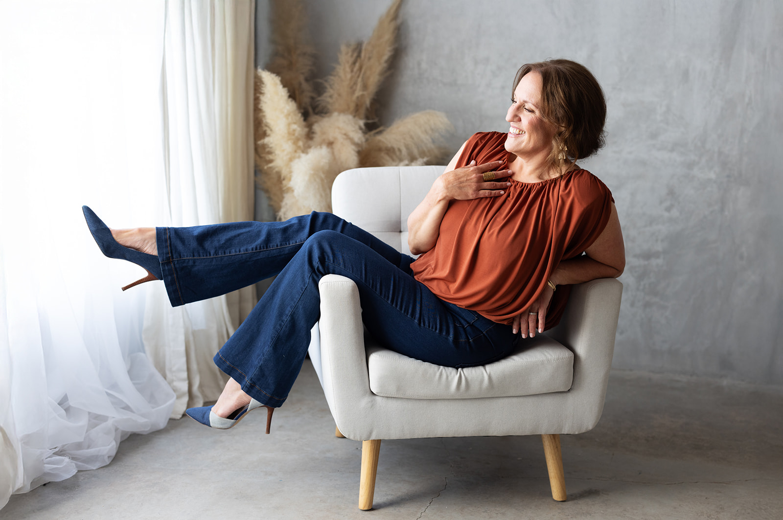 Woman sitting on a white armchair, smiling, with one leg extended, in a bright room with a gray wall and plants.