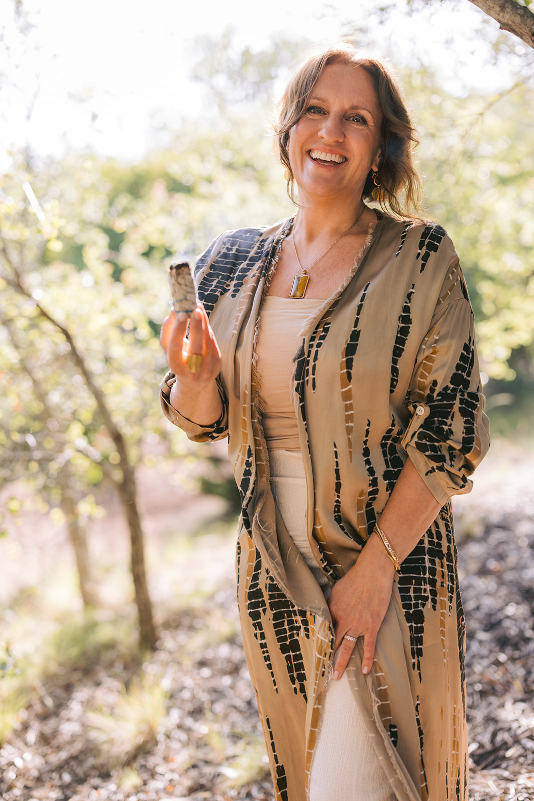 Smiling woman outdoors holding a glass, wearing a patterned jacket and light-colored outfit, trees in background.