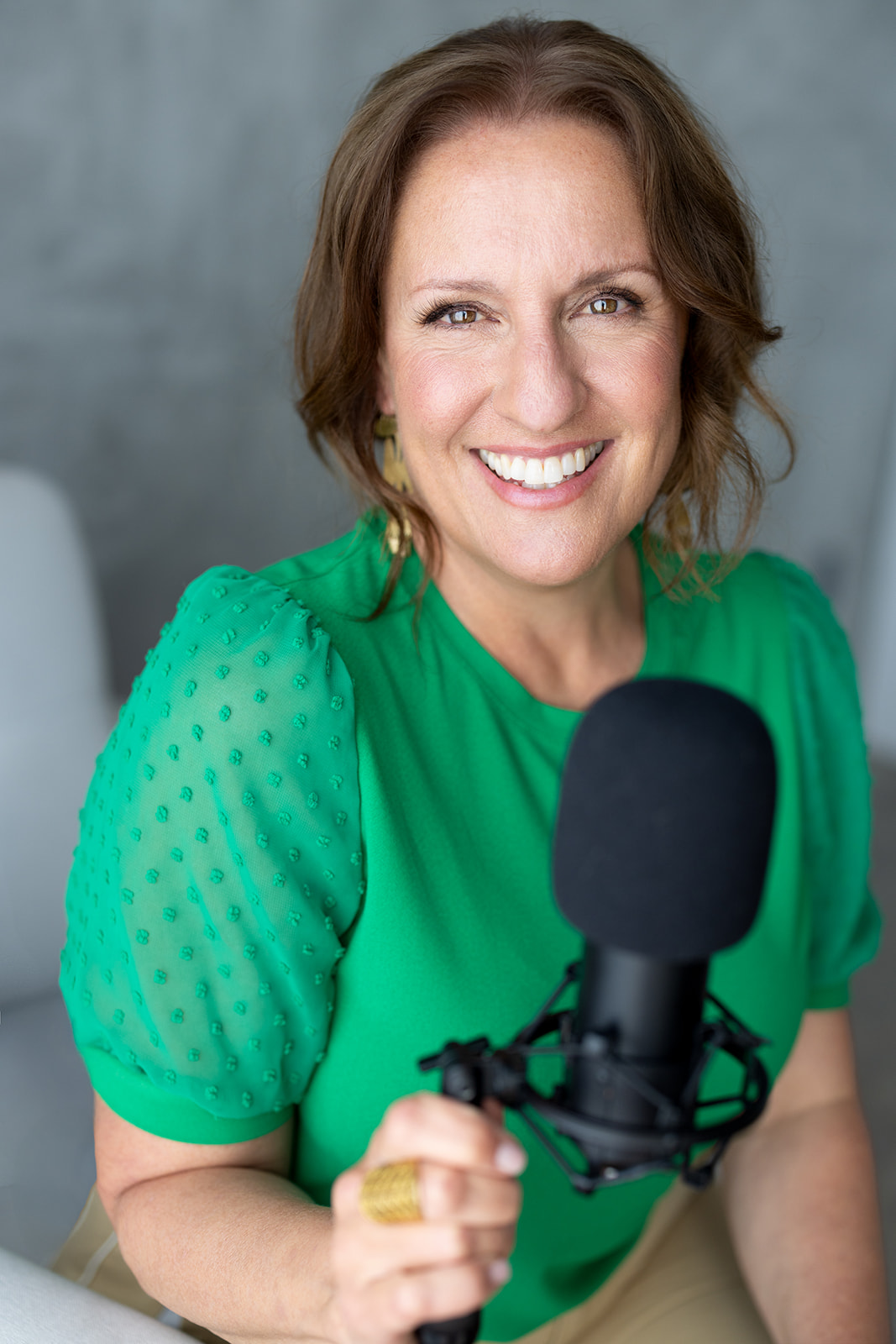 Smiling woman with shoulder-length brown hair wearing a green top, speaking into a microphone.
