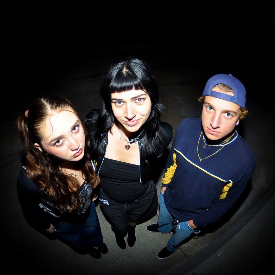 Three young people standing close together, looking up at the camera, with a dark background.
