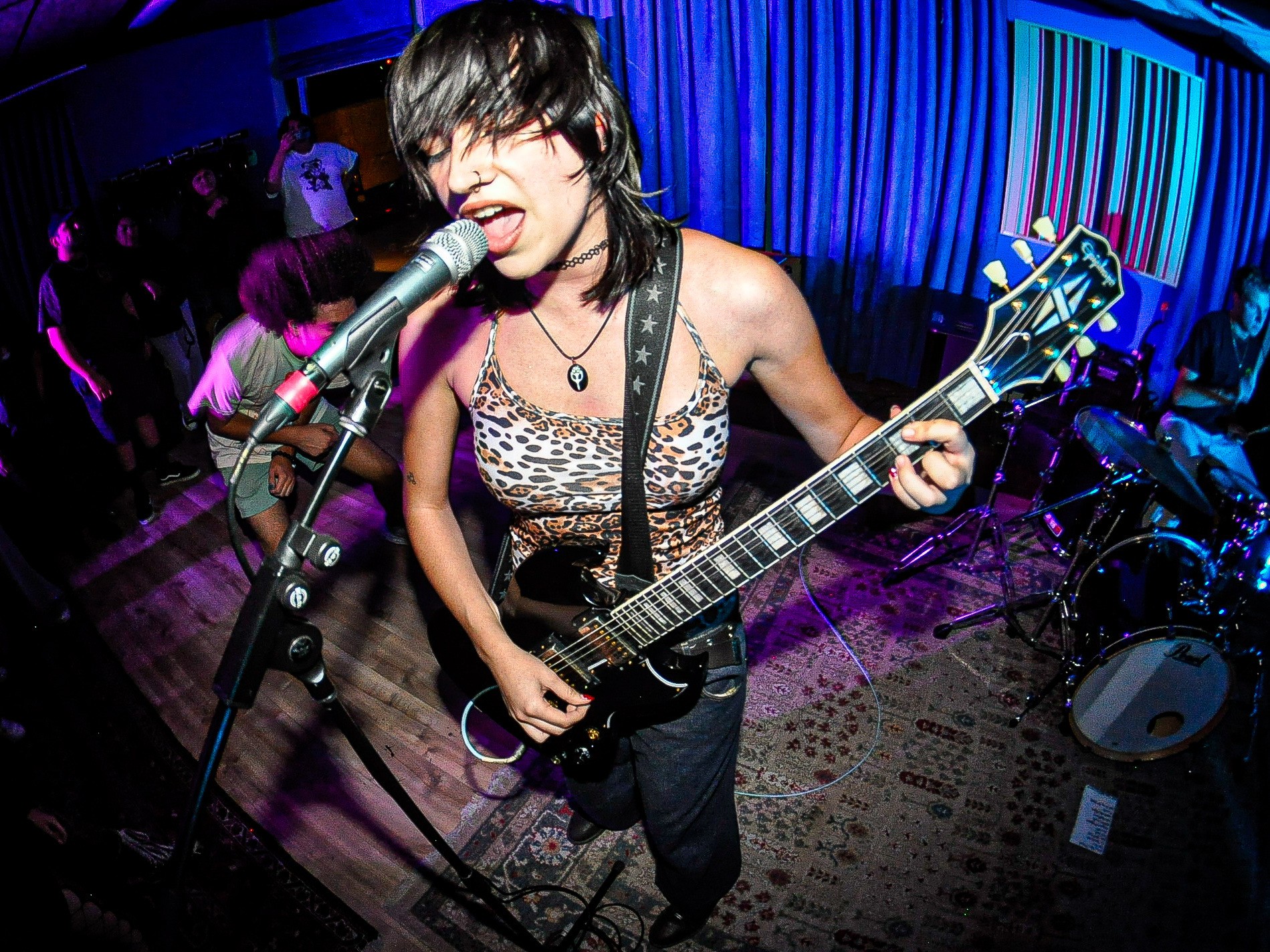 Female musician singing into microphone and playing electric guitar on stage with blue curtains behind her.
