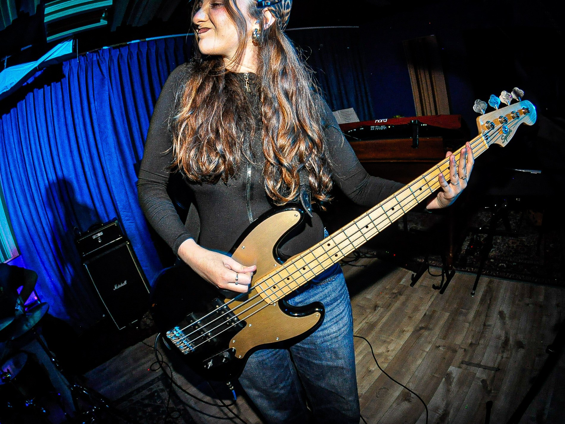 Woman playing bass guitar on stage with blue curtains and musical equipment, smiling and looking to the side.