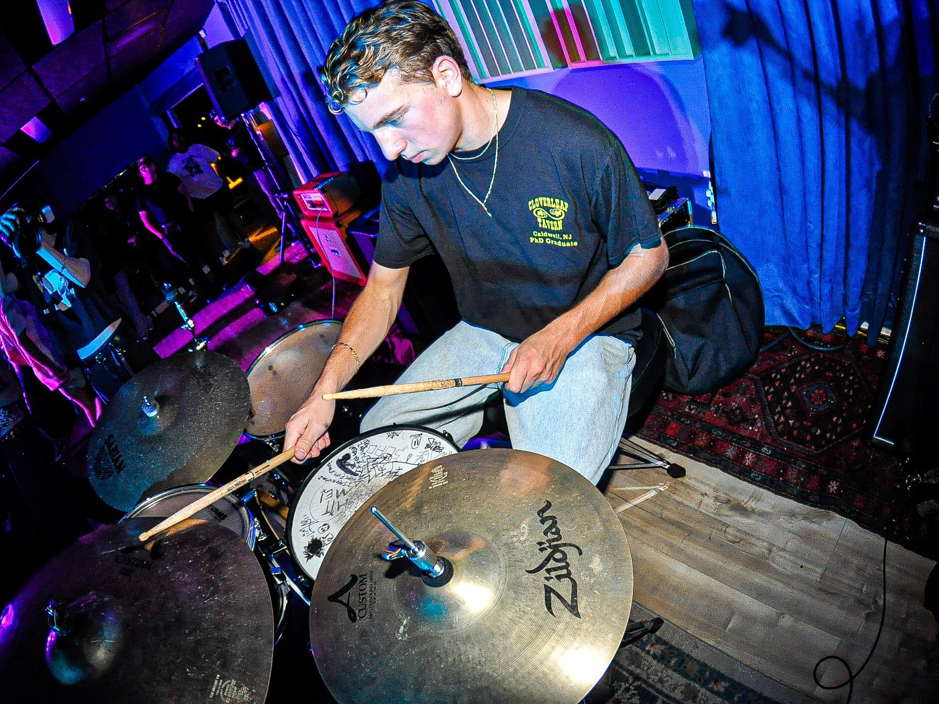 Young man playing cymbals at a music event with colorful lighting, wearing a black t-shirt and jeans.