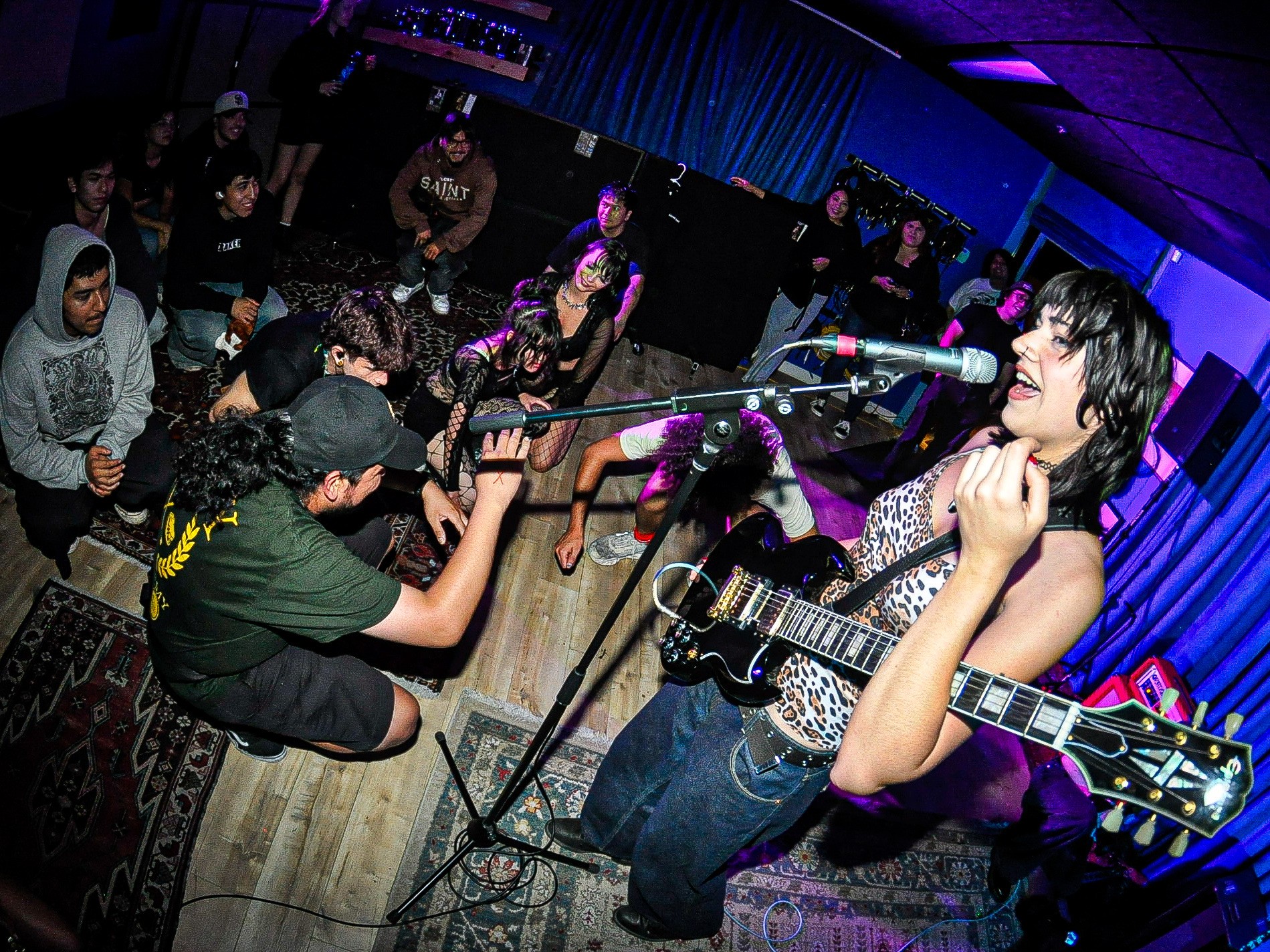 Female musician singing and playing guitar on stage with audience watching, blue stage curtains in background.