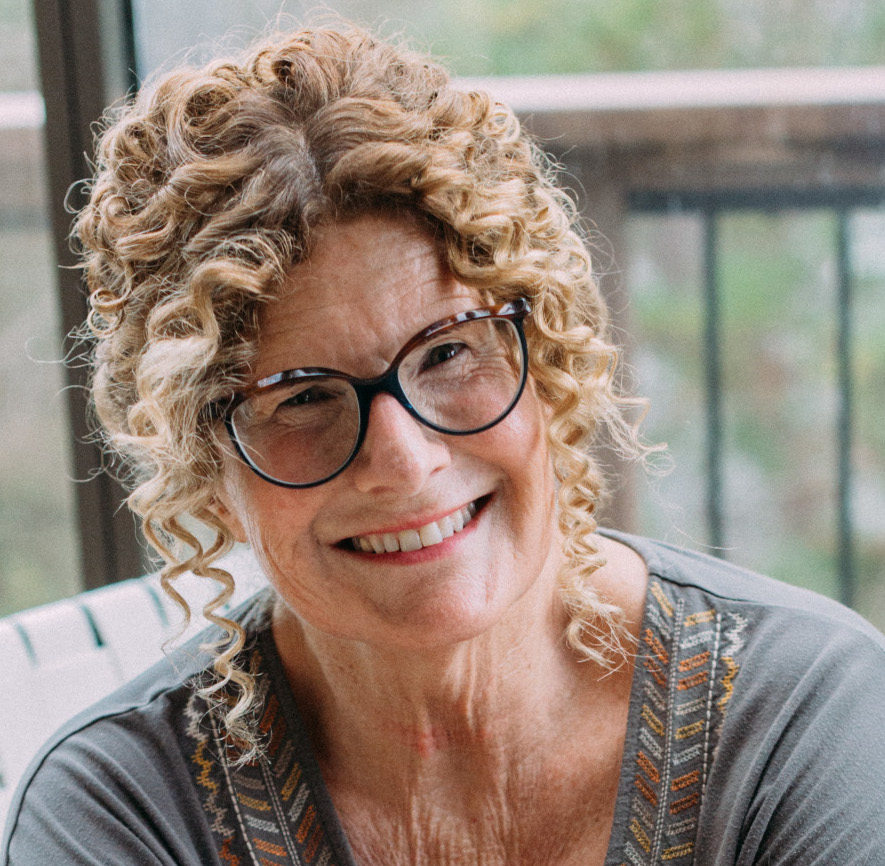 Smiling woman with curly hair and glasses indoors near a window.