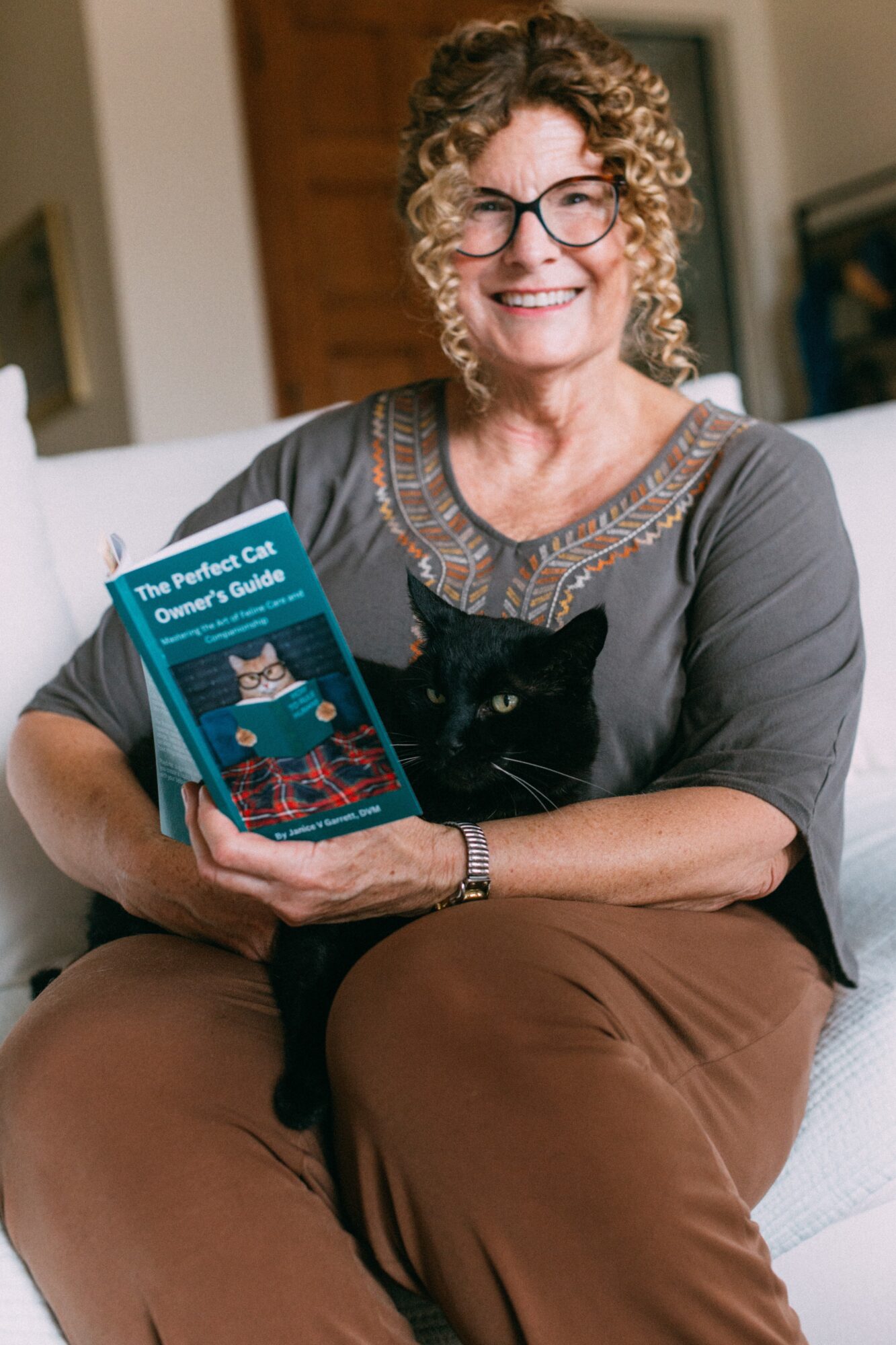 Woman with curly hair and glasses holding a book and a black cat, sitting on a couch.