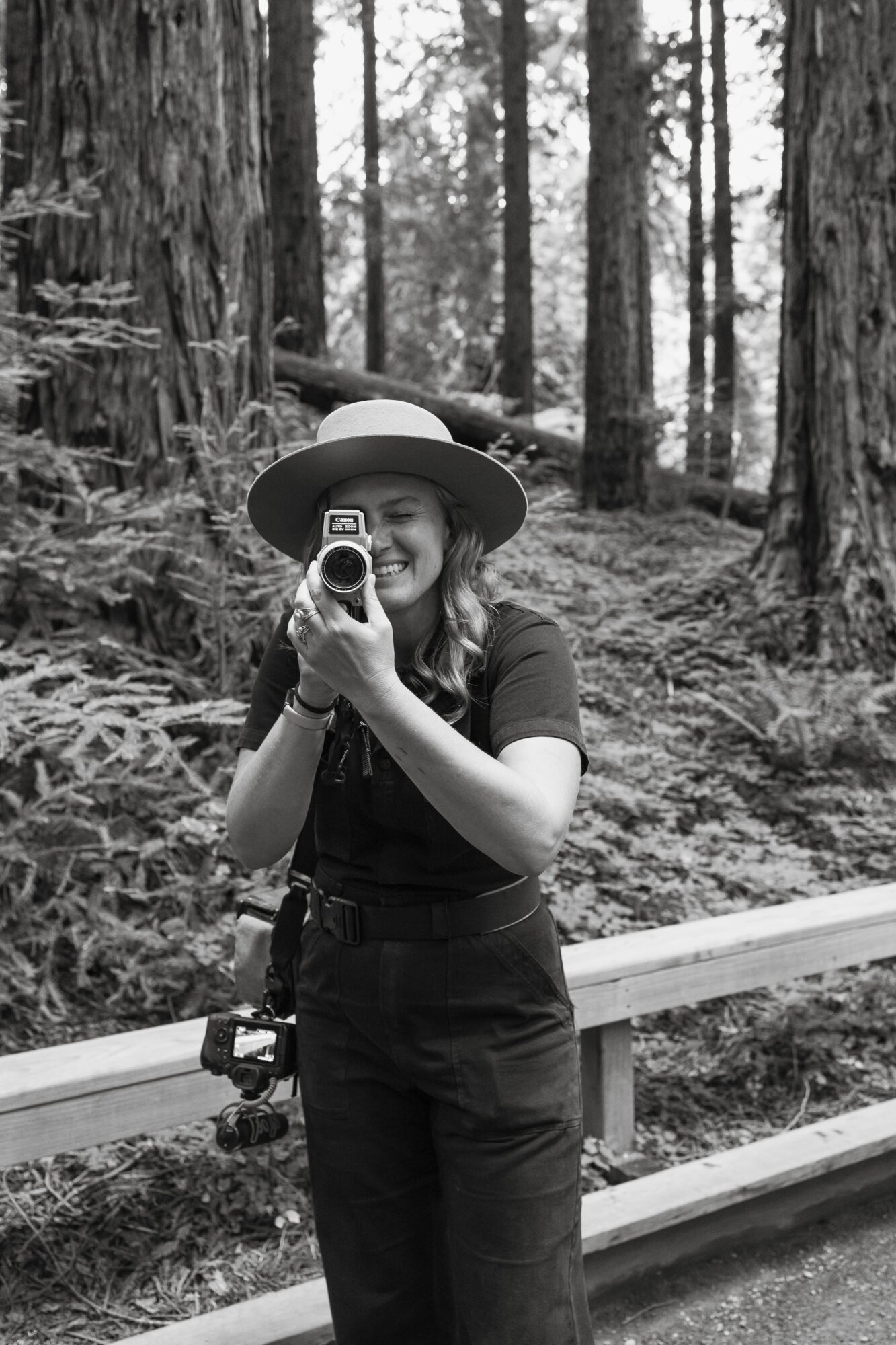 Person wearing a hat and smiling, holding a camera, standing outdoors in a forested area with trees and a railing.