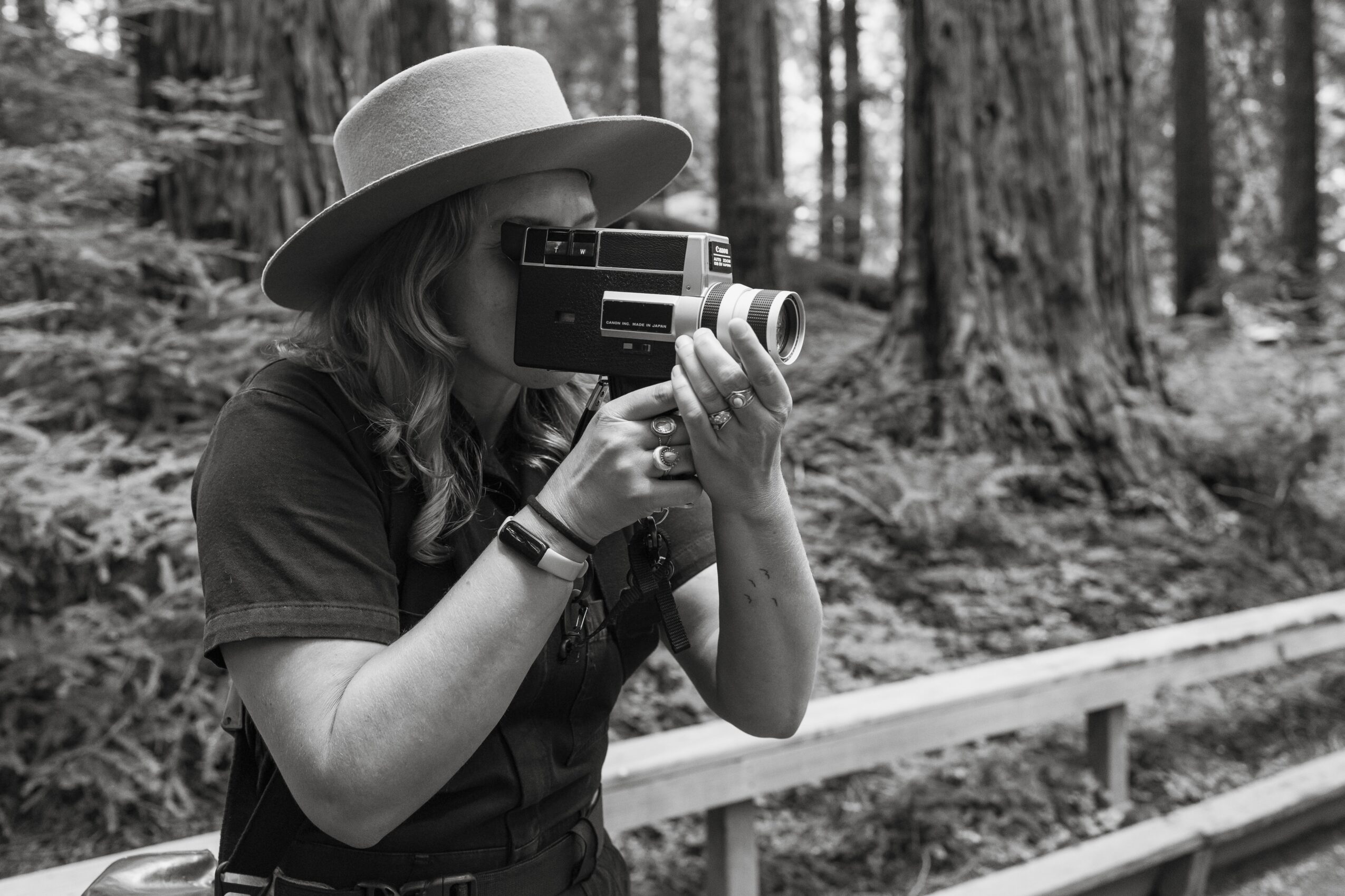 Person wearing a hat taking a photo with a camera outdoors in a forest, black and white image.
