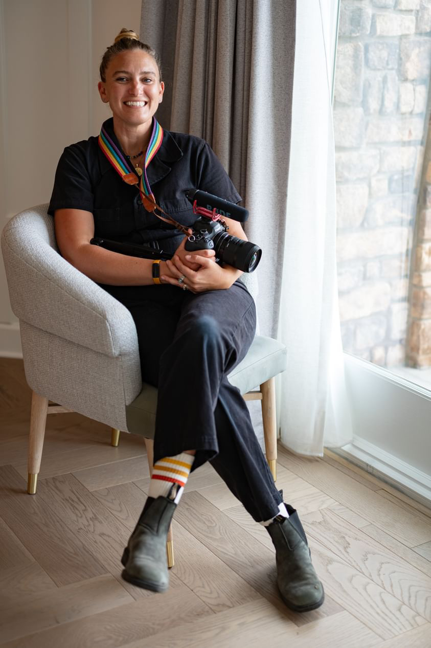 Person sitting on a chair holding a camera, smiling, near a window with curtains, in a room with wooden floor.