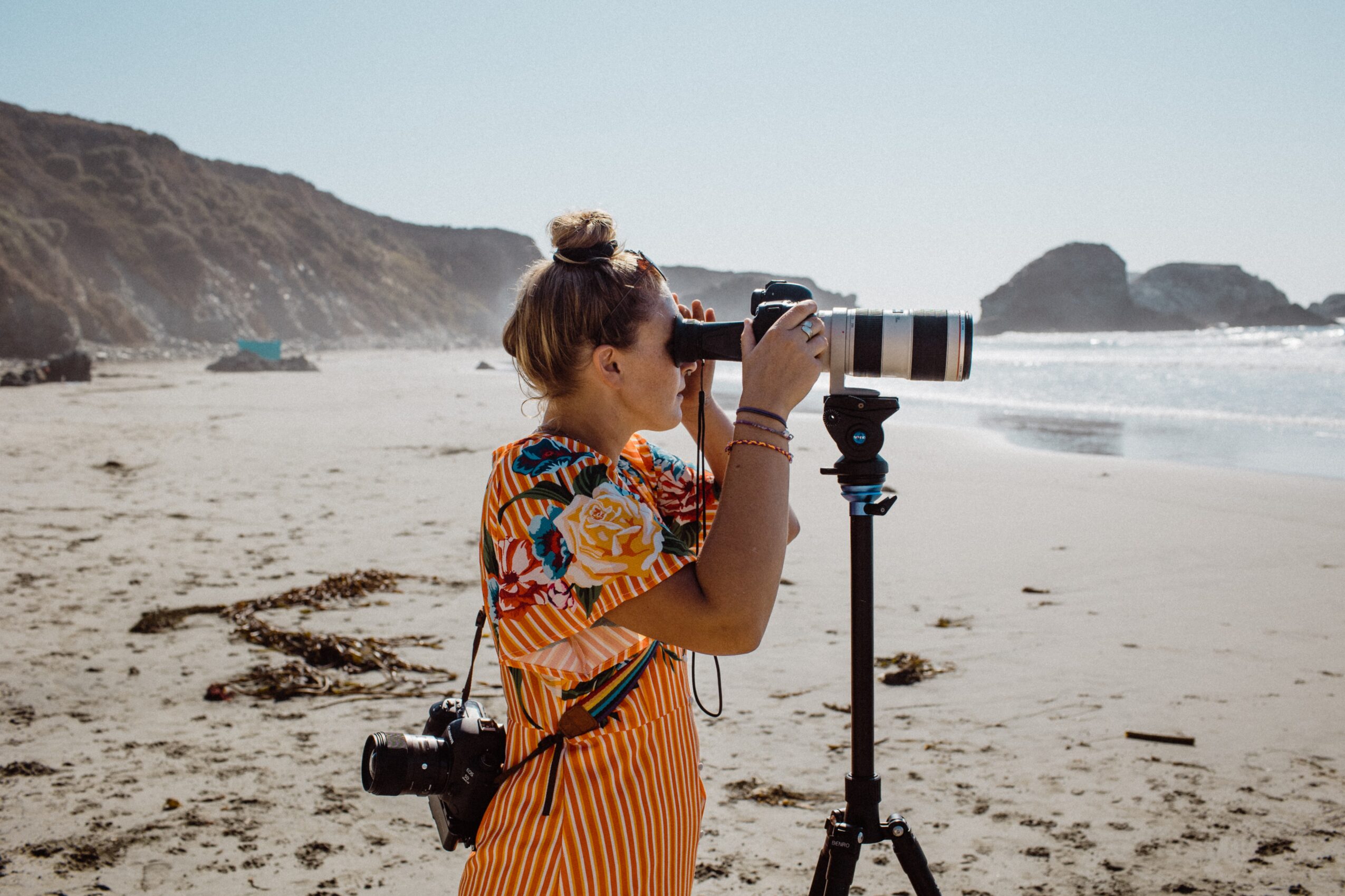 Person with a camera on a tripod at the beach, looking through a telephoto lens, with a camera hanging from their shoulder.