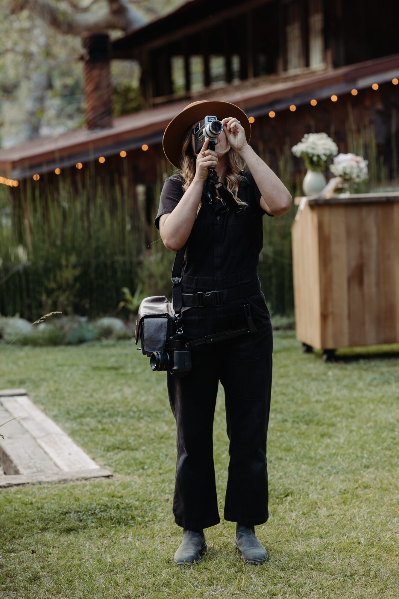 Person standing outdoors taking a photo with a camera, wearing a hat and black clothing, with a wooden structure and flowers in background.