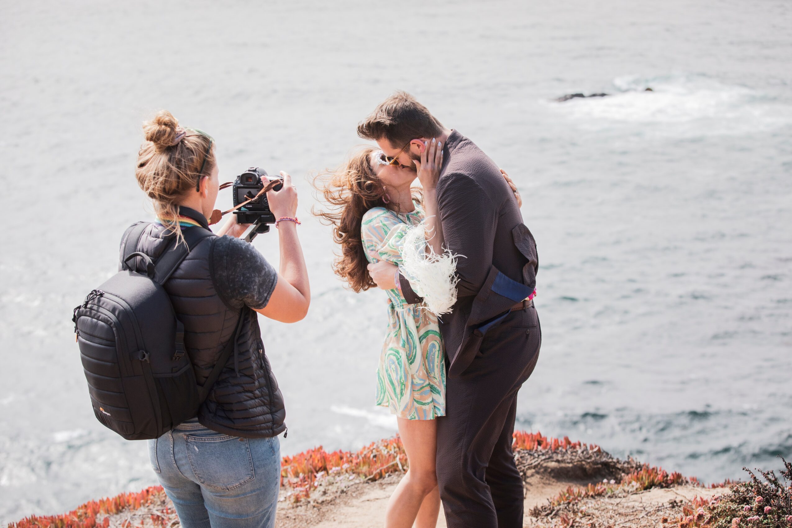 A person with a backpack takes a photo of a couple kissing near the water's edge, with a rocky shoreline and ocean in the background.
