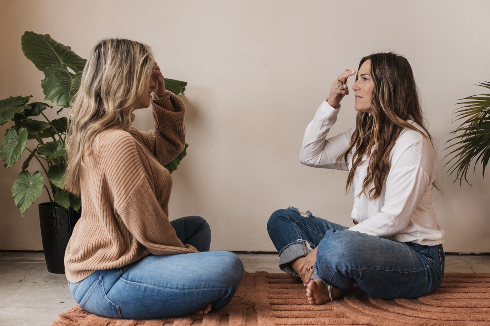 Two women sit on the floor facing each other, engaging in conversation, with plants in the background.