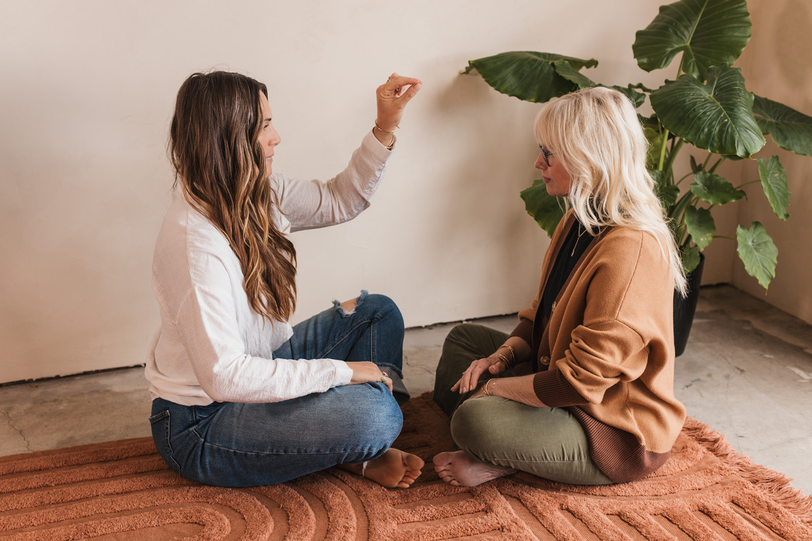 Two women sit on a rug facing each other, engaging in a conversation indoors with a large plant nearby.