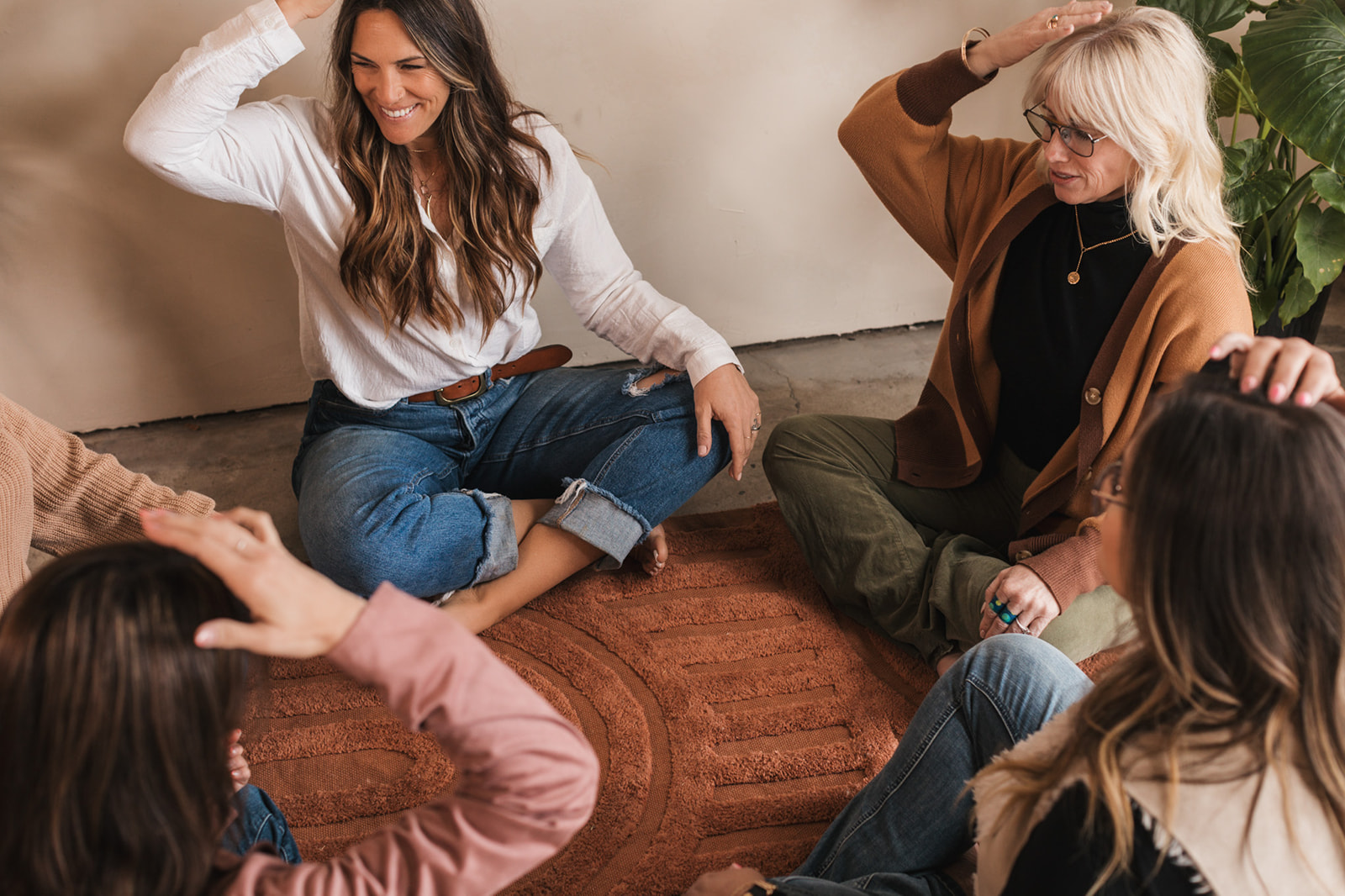 Four women sitting on a rug, smiling and engaging in conversation indoors.