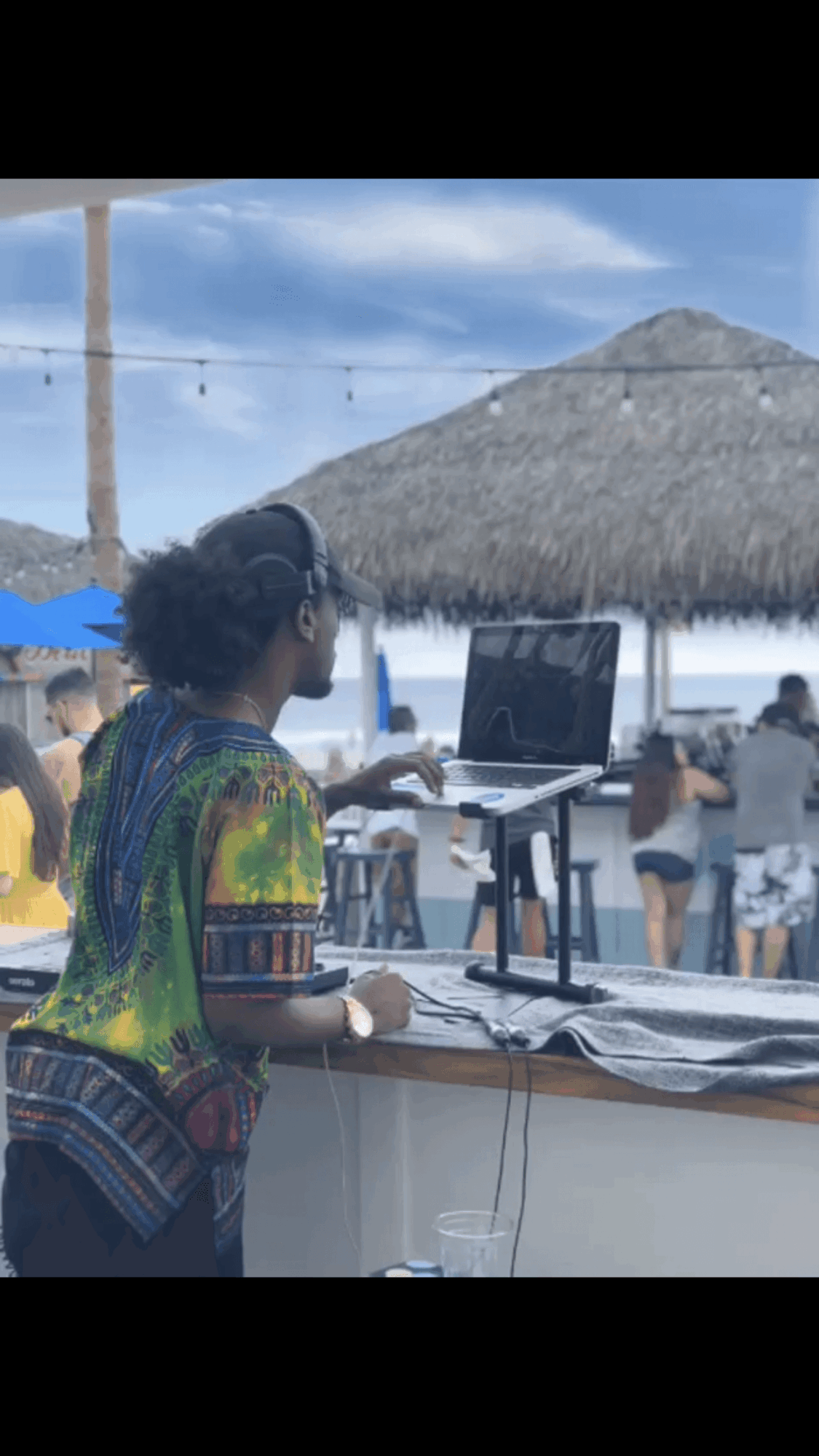 Person working on a laptop at an outdoor beachside venue with thatched roof, people in the background, and a blue sky.