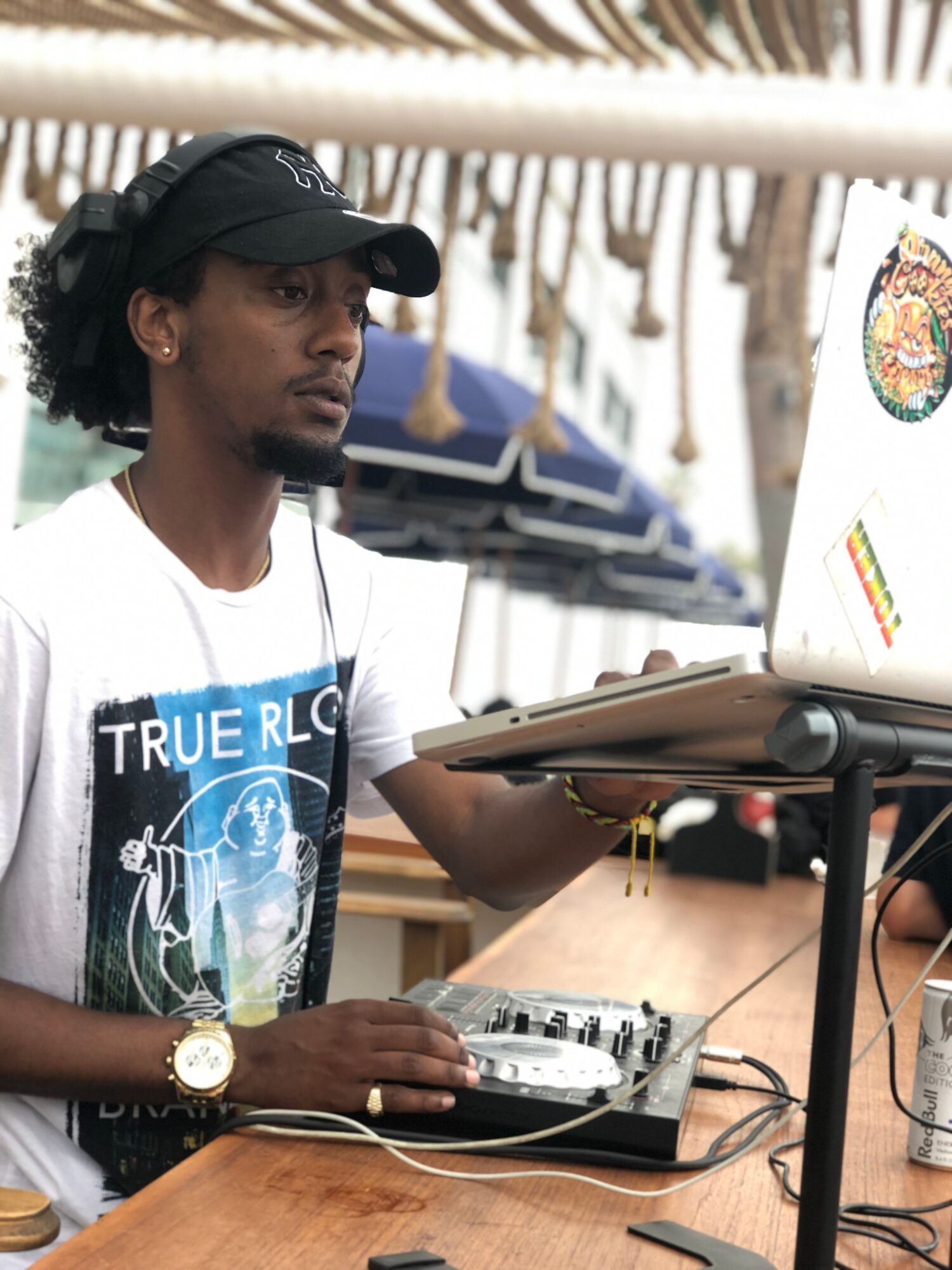 Person wearing a black cap and white T-shirt operating DJ equipment outdoors, with a laptop and a drink on the table.