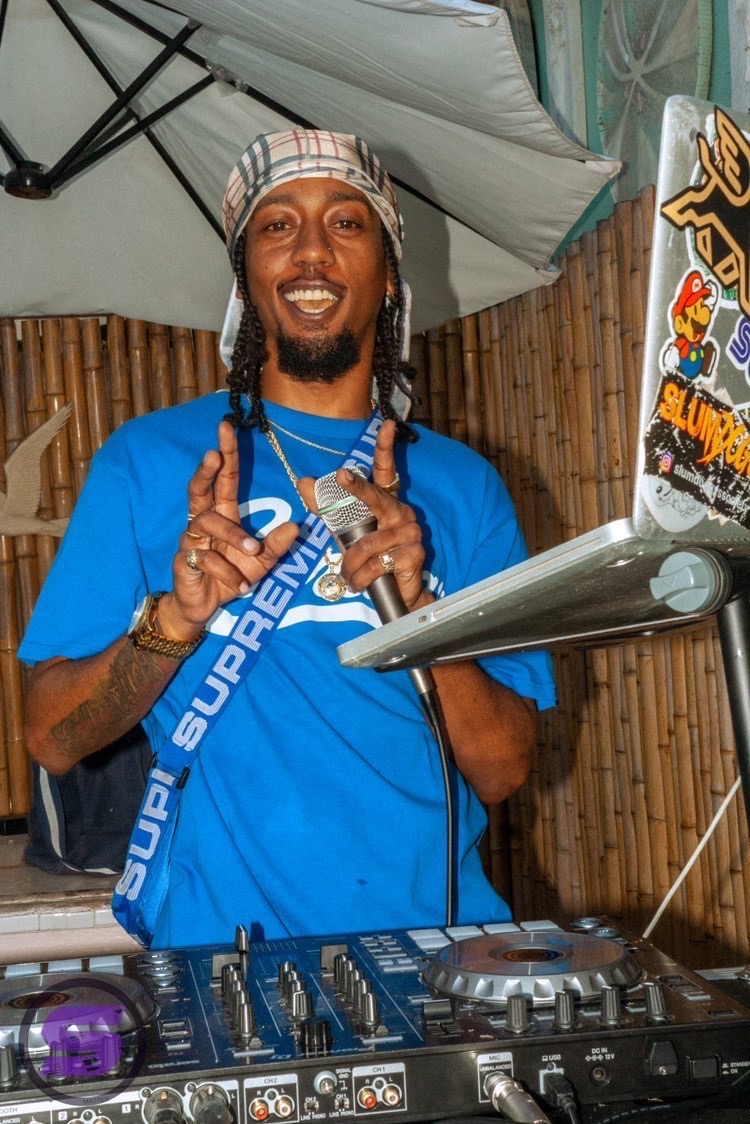 DJ with dreadlocks, wearing a blue shirt and a patterned headscarf, smiling and pointing at the camera, with DJ equipment in front.